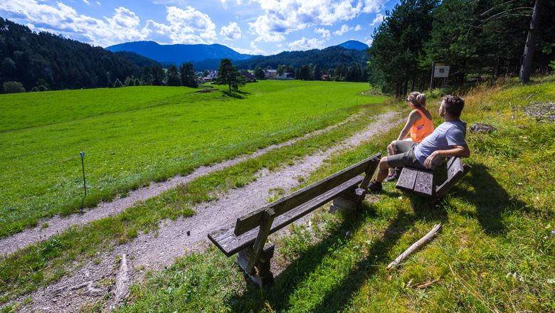 Two people sit on a bench and look out over a green meadow with mountains in the background.