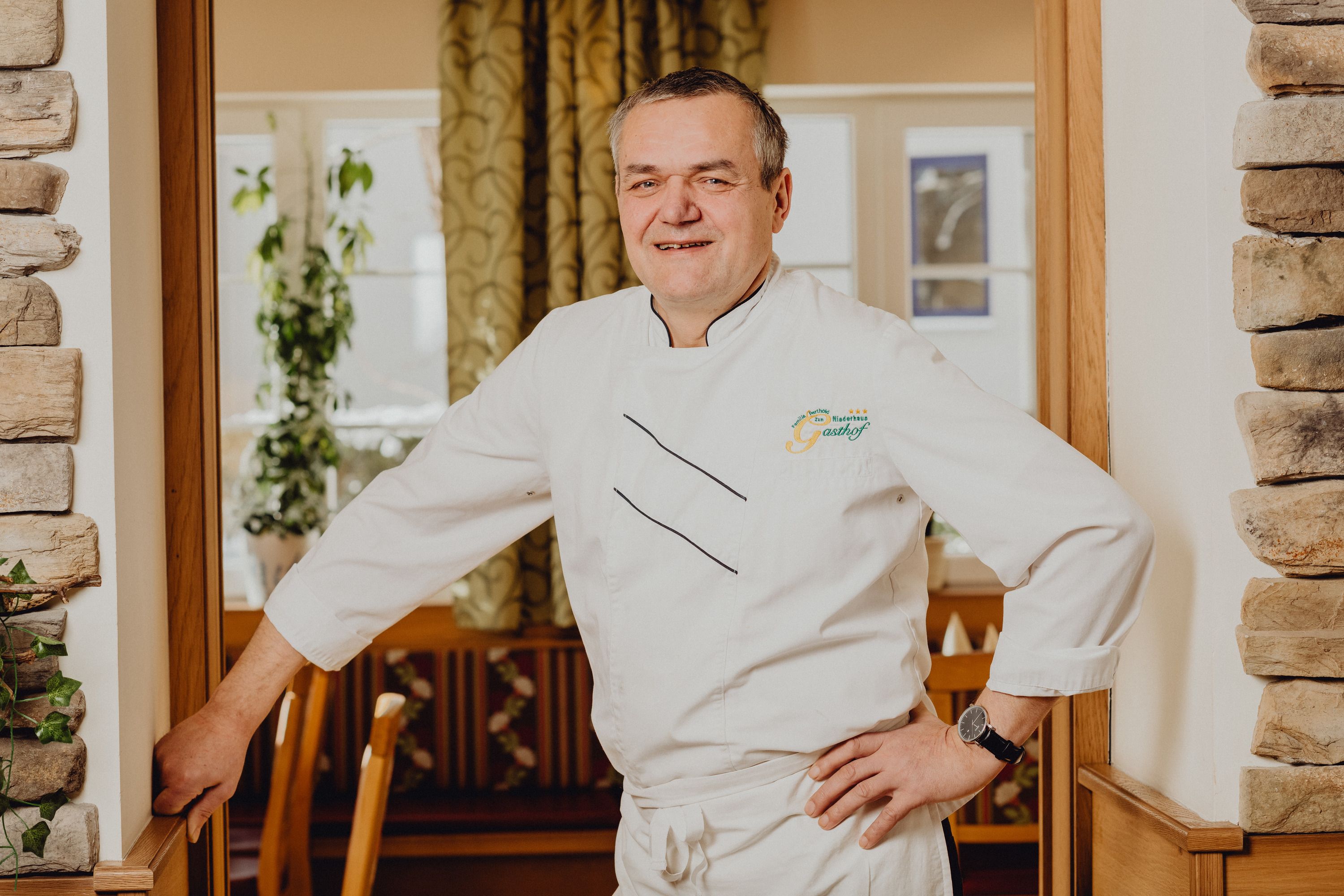 A man in a white chef's uniform stands smiling in a restaurant.