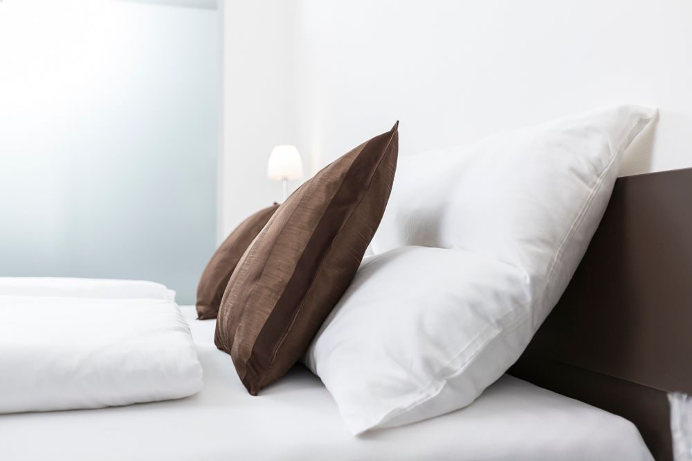 A bed with white cushions and brown decorative pillows, a lamp in the background.