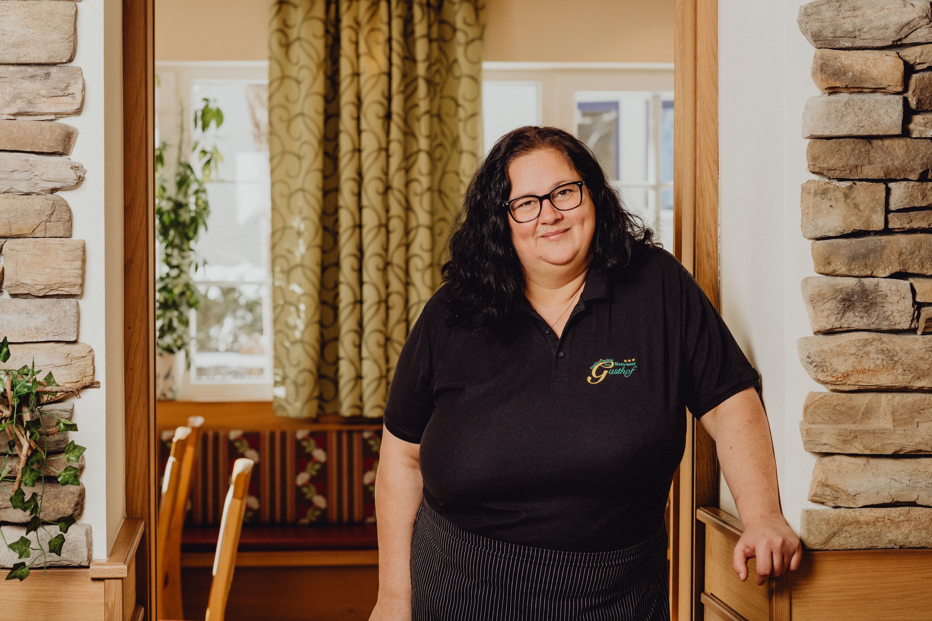 A woman with glasses stands in a cozy inn with stone walls and curtains.