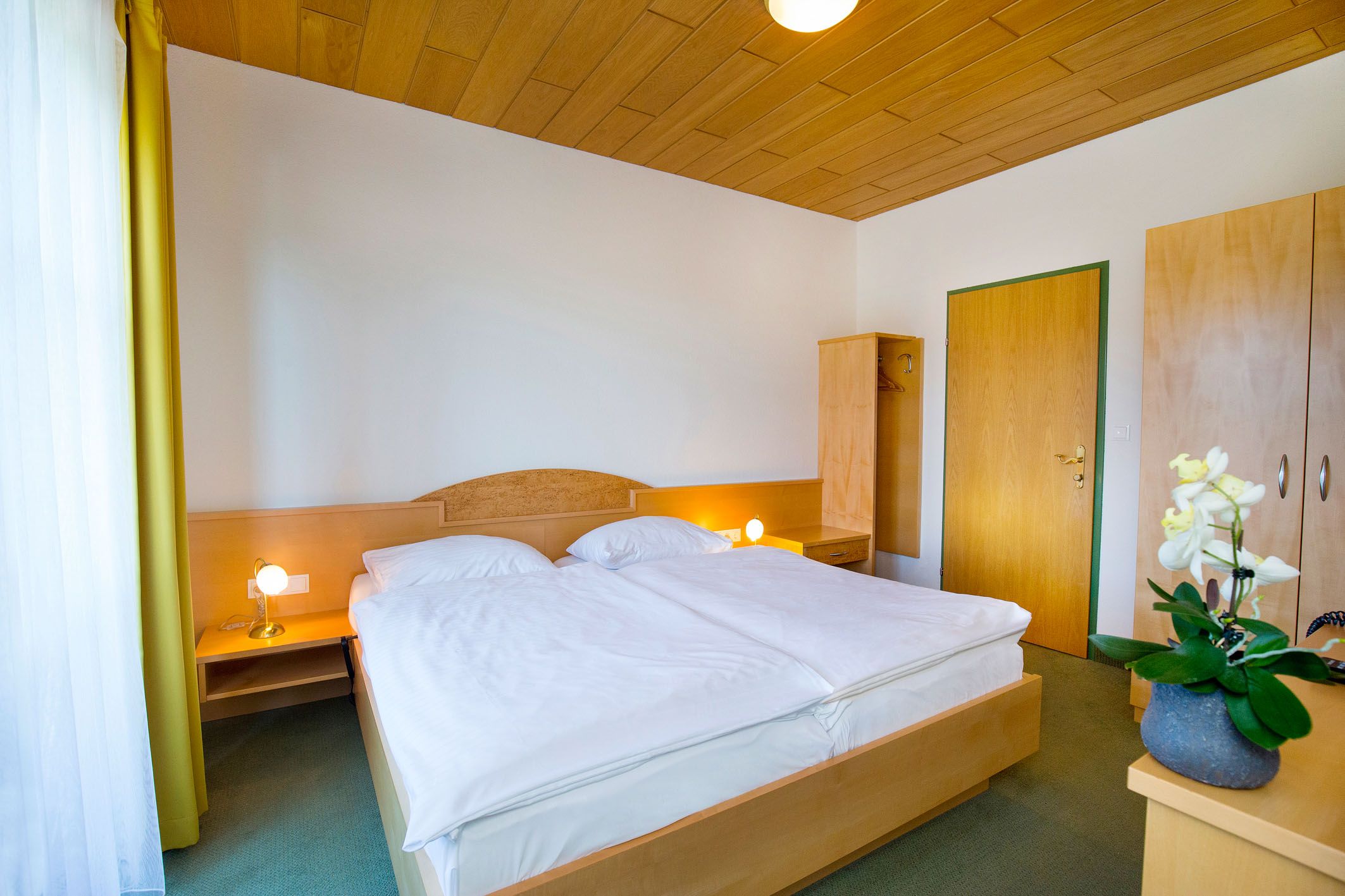 A simply furnished hotel room with a double bed, wooden ceiling, wardrobe and bedside lamps.