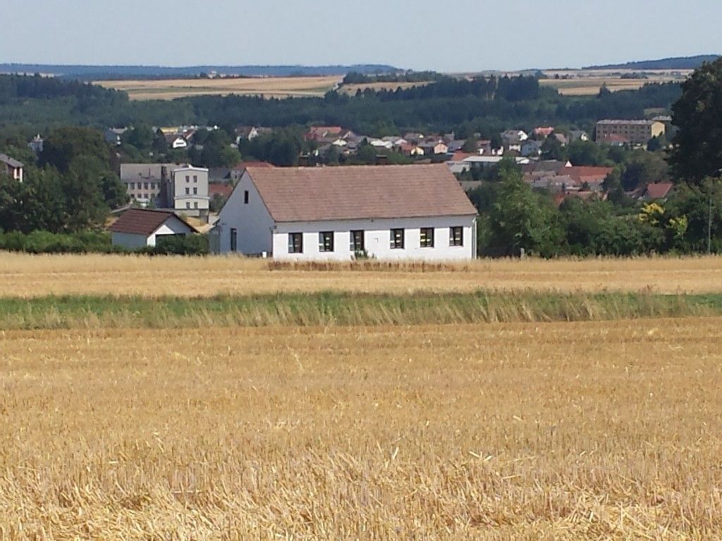 A white house with a red roof stands in a field, with a town and woods in the background.