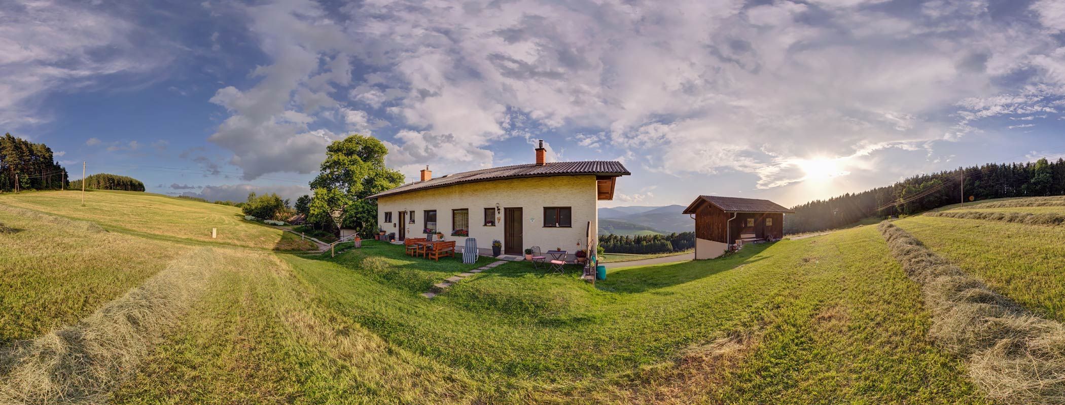 Panoramic view of a house on a meadow with trees and mountains in the background at sunset.