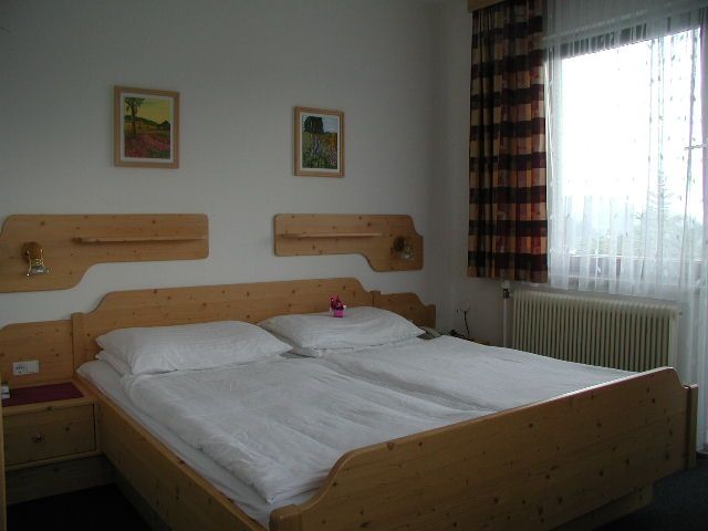 A simply furnished hotel room with a wooden double bed, two pictures on the wall and a window with curtains.