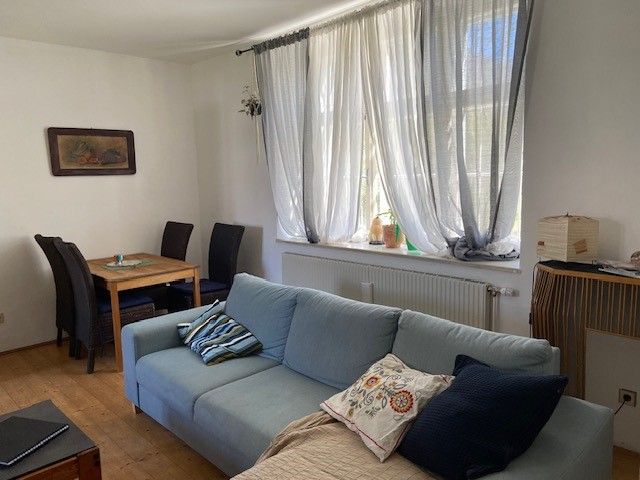A living room with a blue sofa, a wooden table with chairs and a window with white curtains.