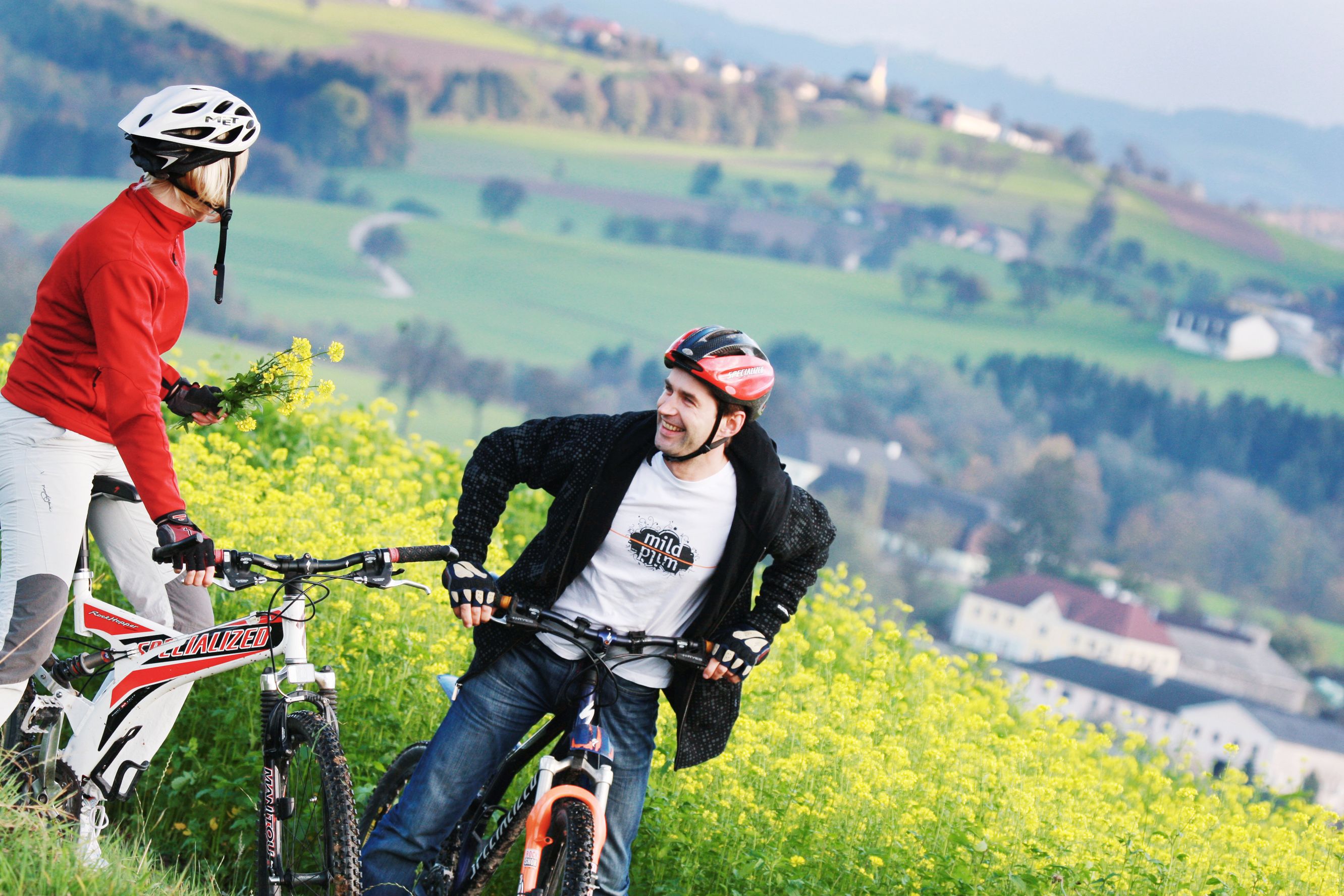 Two cyclists with helmets are standing in a flowering field with a green landscape in the background.