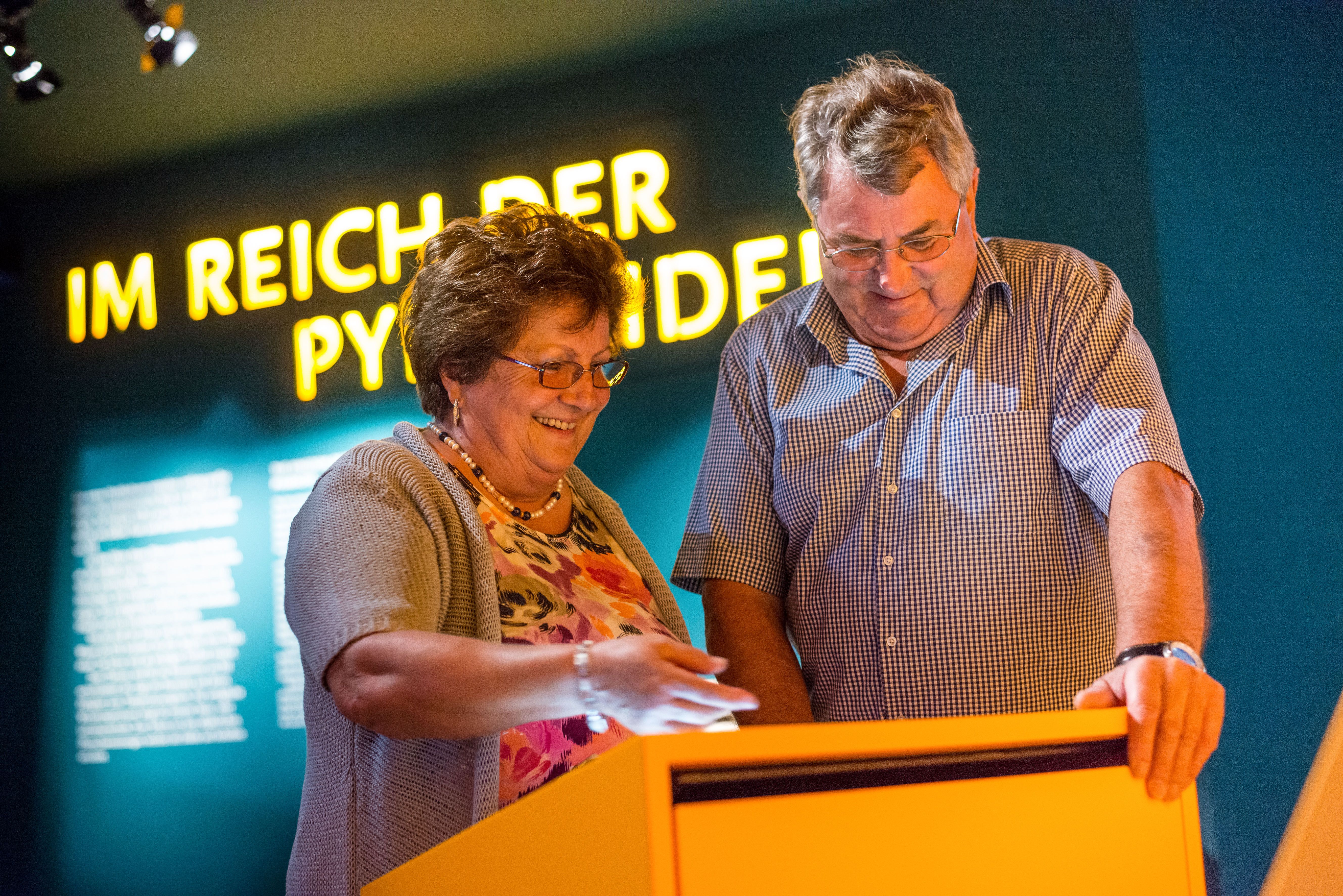 Two people look at an interactive display in a museum with the inscription 'In the realm of the pyramids' in the background.