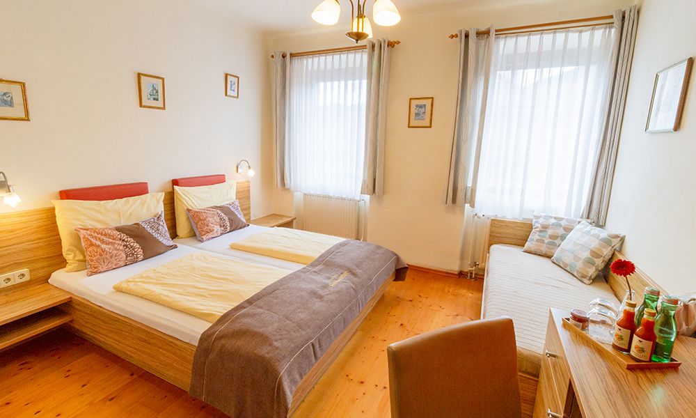 A cozy guest room with double bed, day bed, wooden furniture and decorative cushions. Bright curtains and pictures on the walls.