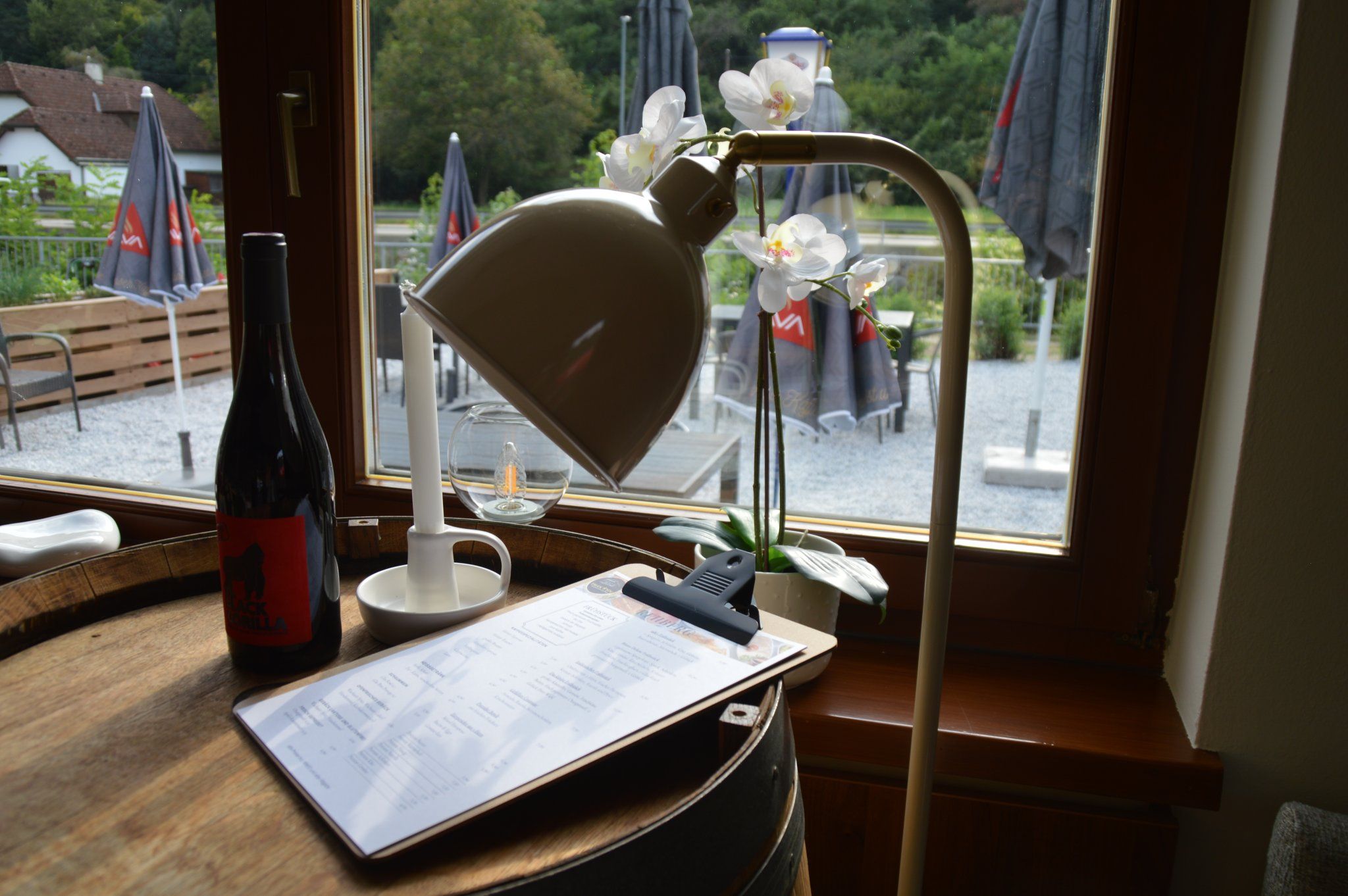 Interior view of a bistro with wine bottle, lamp and menu on a barrel, view outside onto terrace with parasols.