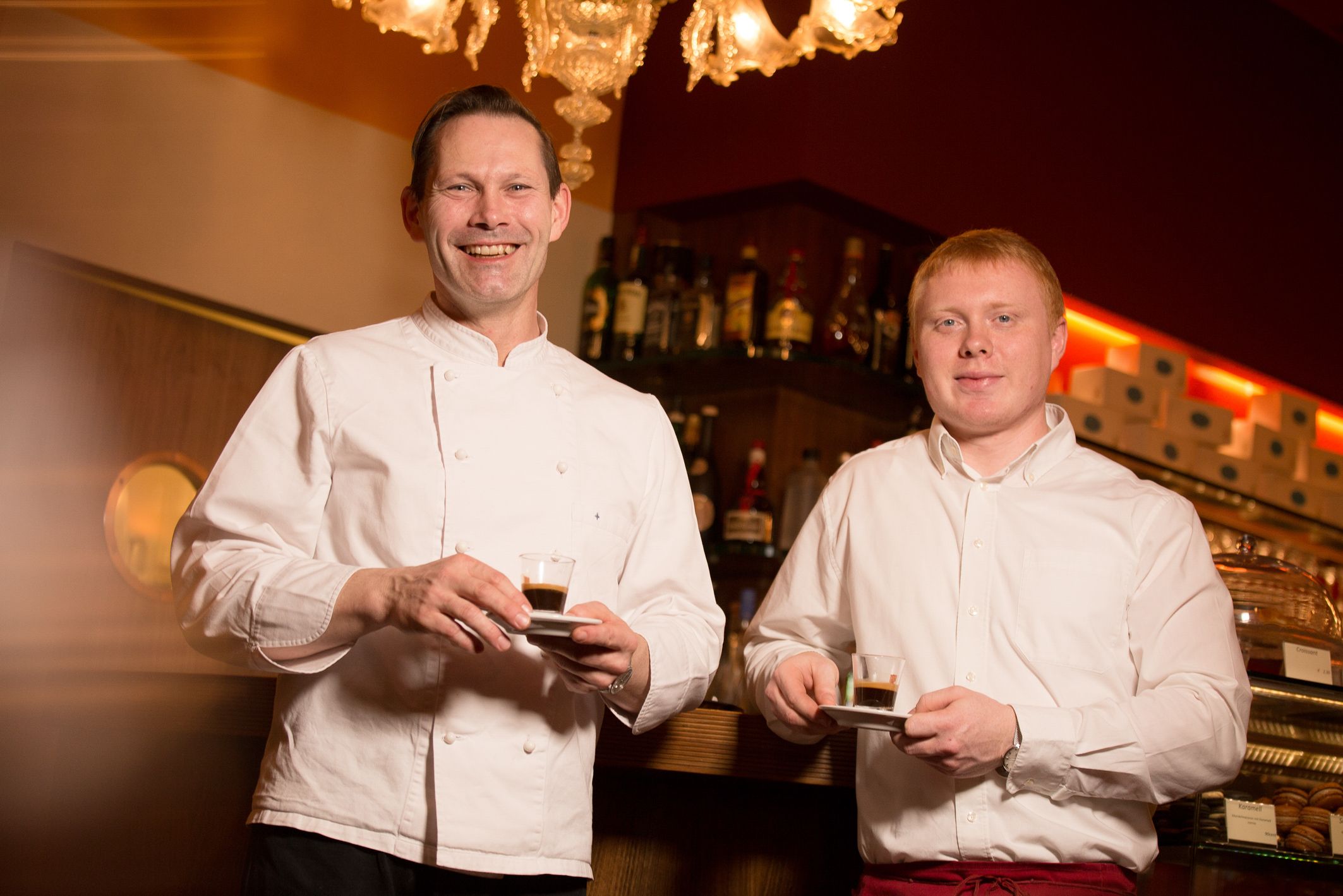 Two men in a café holding espresso cups and smiling at the camera.