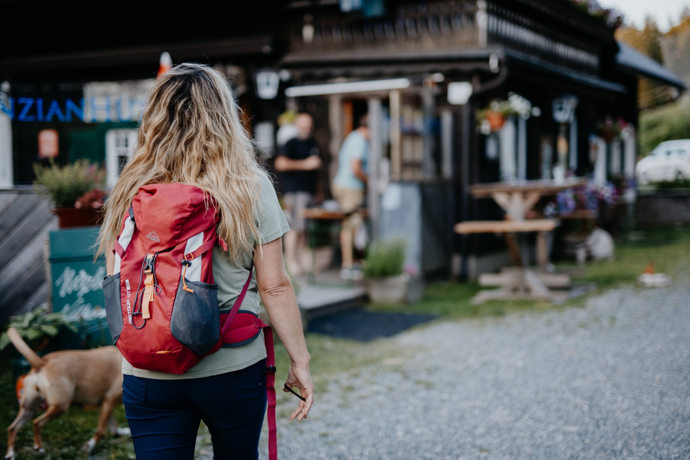 Woman with a red rucksack and dogs walking towards a mountain hut.