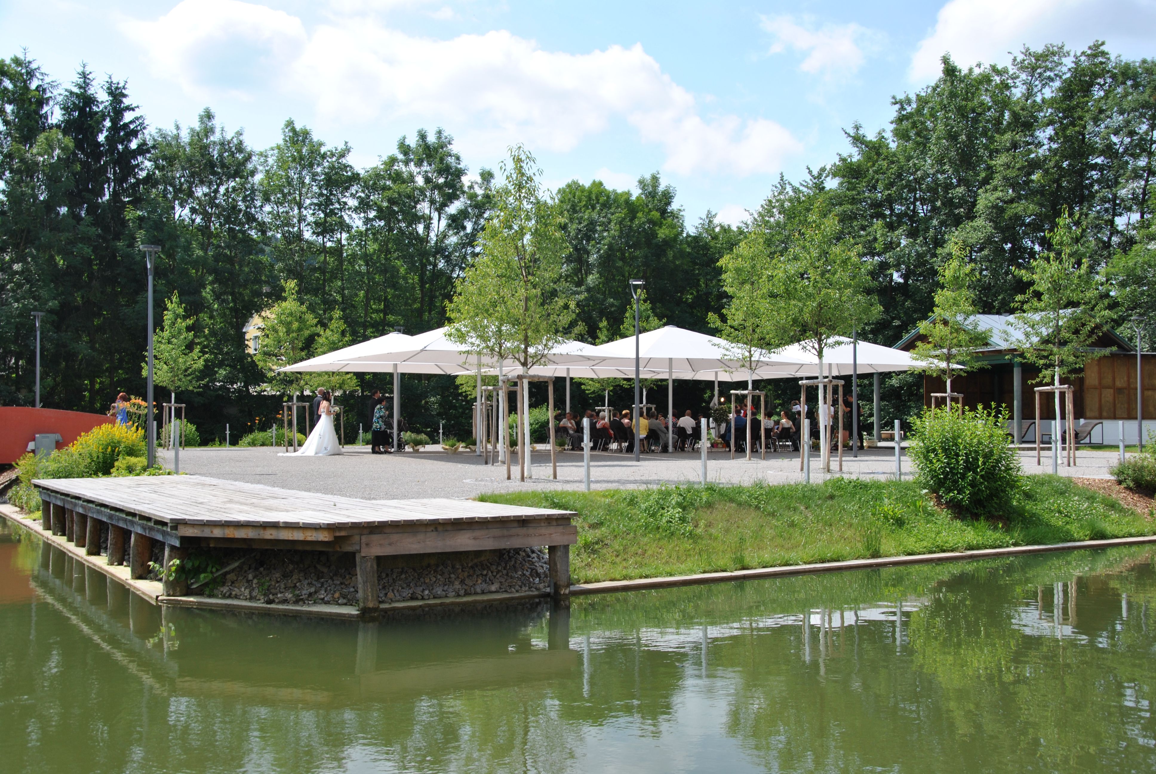 A wedding couple stands in front of a pavilion with guests, surrounded by trees and a pond.