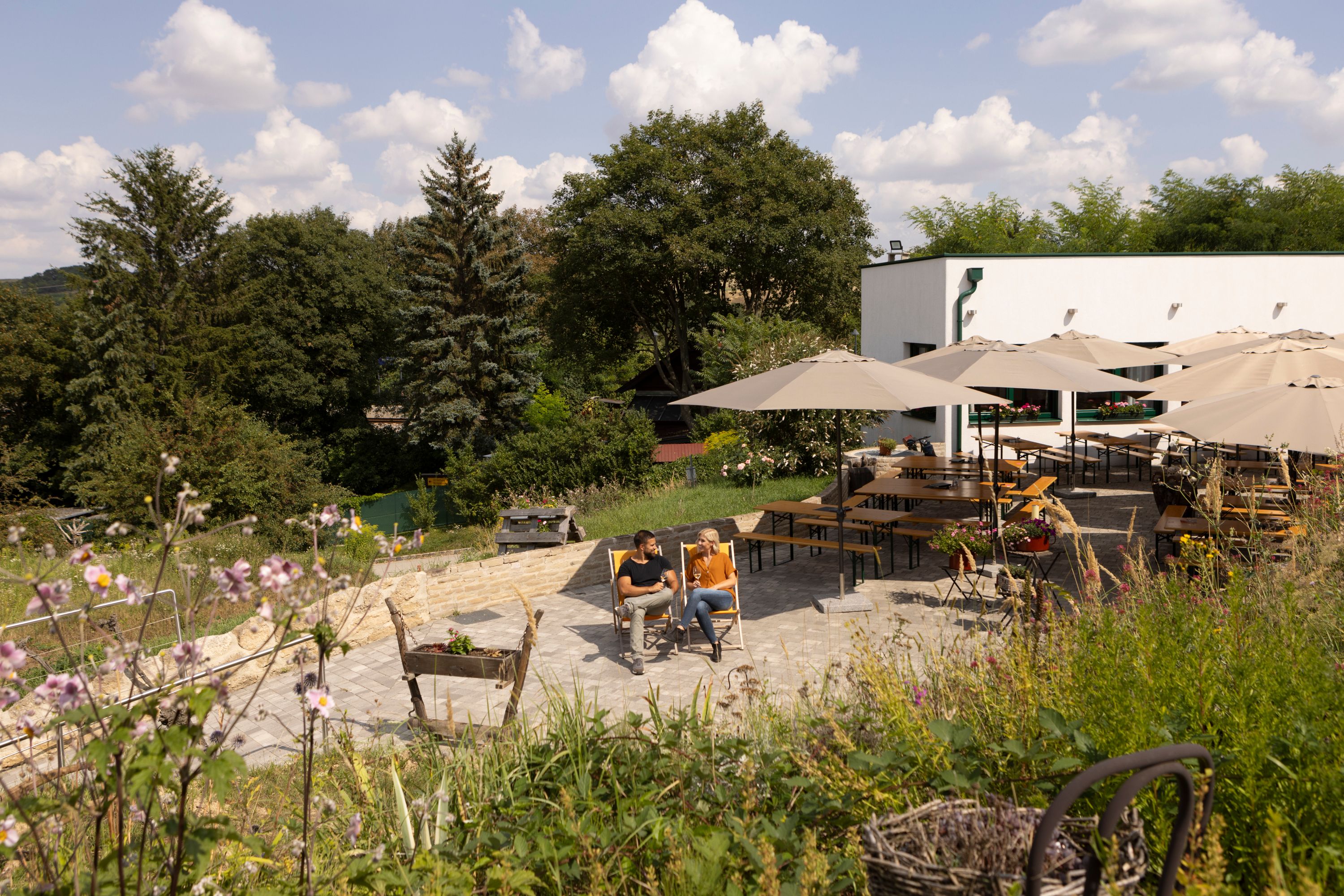 Two people sit on deckchairs in a garden with parasols and tables, surrounded by trees and flowers.