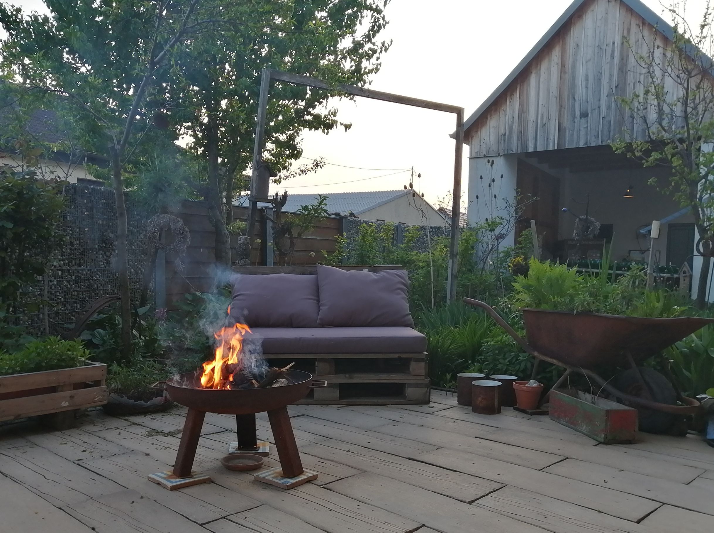 A cozy courtyard with a fireplace, a sofa made from pallets and an old wheelbarrow. A wooden shed and green plants in the background.