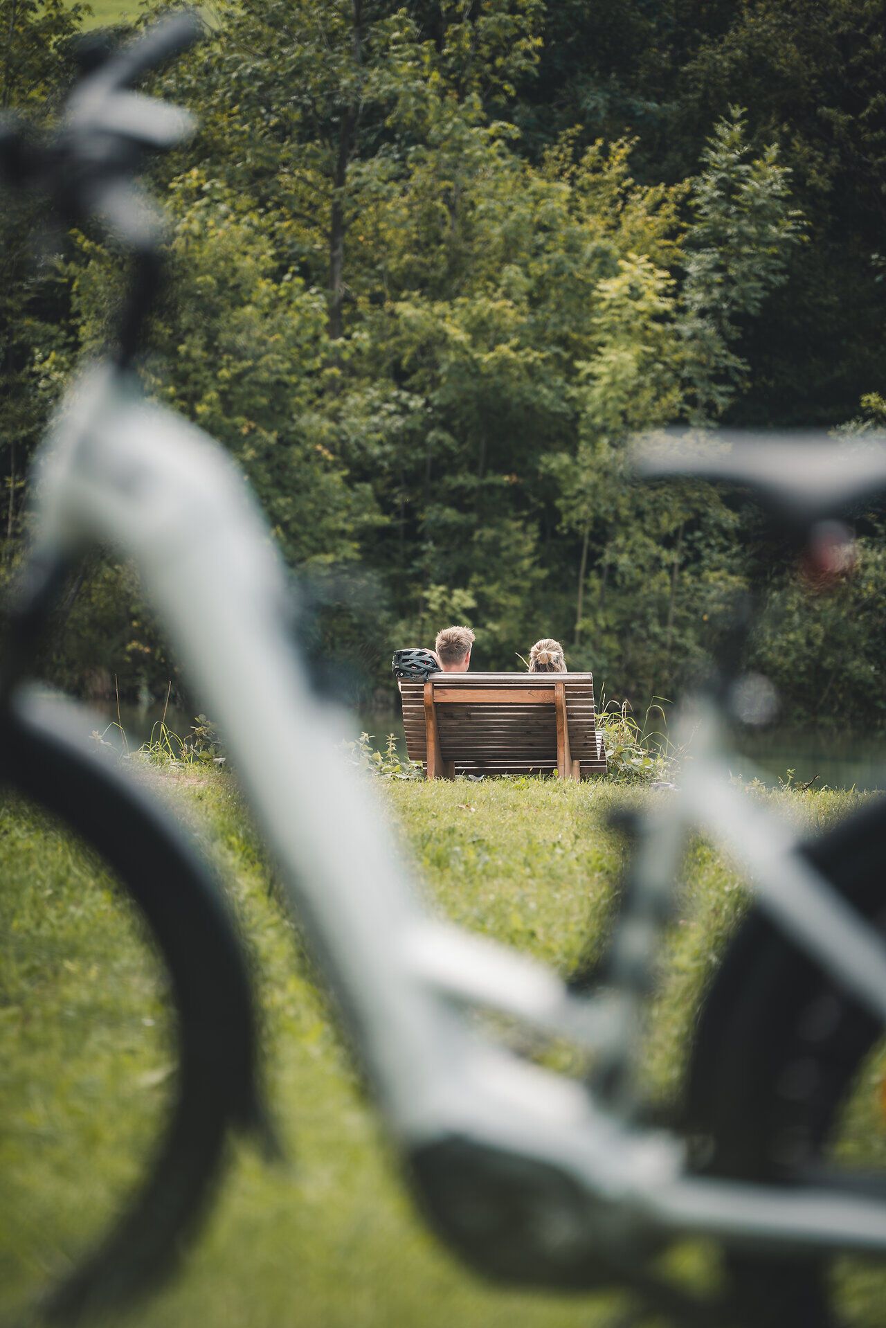 Im Vordergrund steht ein Fahrrad, dahinter entspannen im Fokus zwei Personen auf einer Liege aus Holz mit Blick auf einen Fluss und die Pflanzen dahinter.