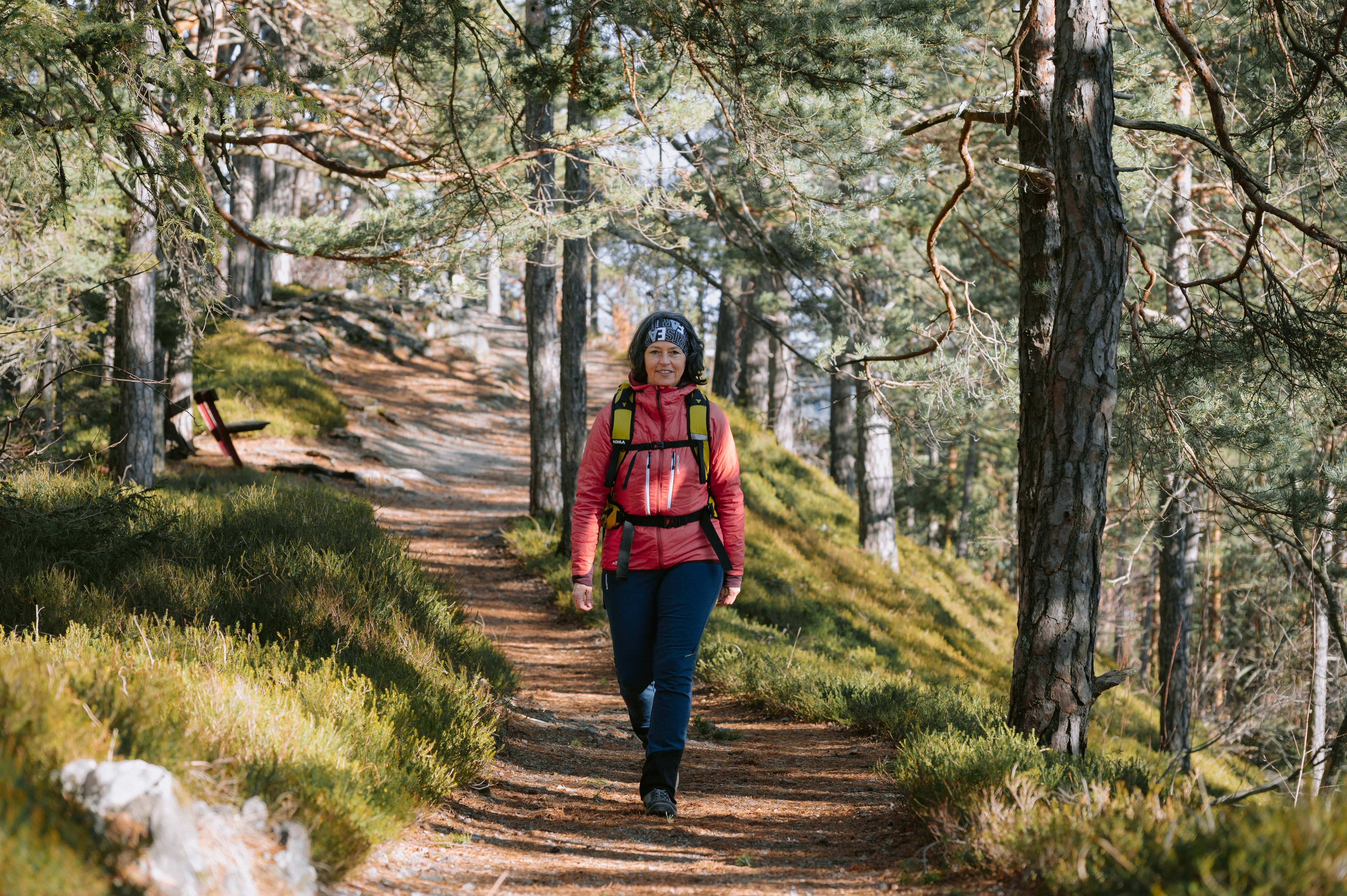 A woman walks along a forest path, surrounded by trees and sunlight.