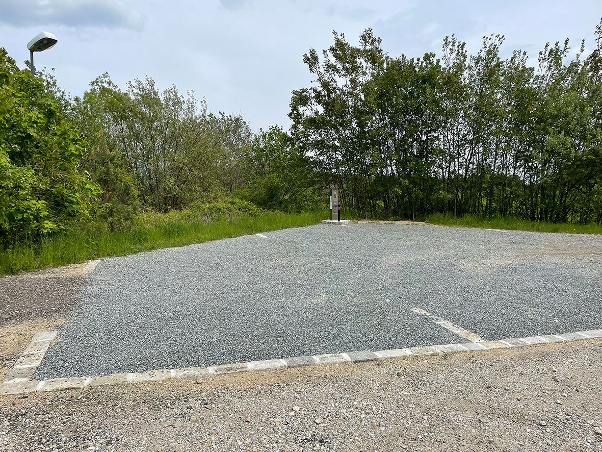 An empty camper van site with a gravel floor, surrounded by trees and a lantern.