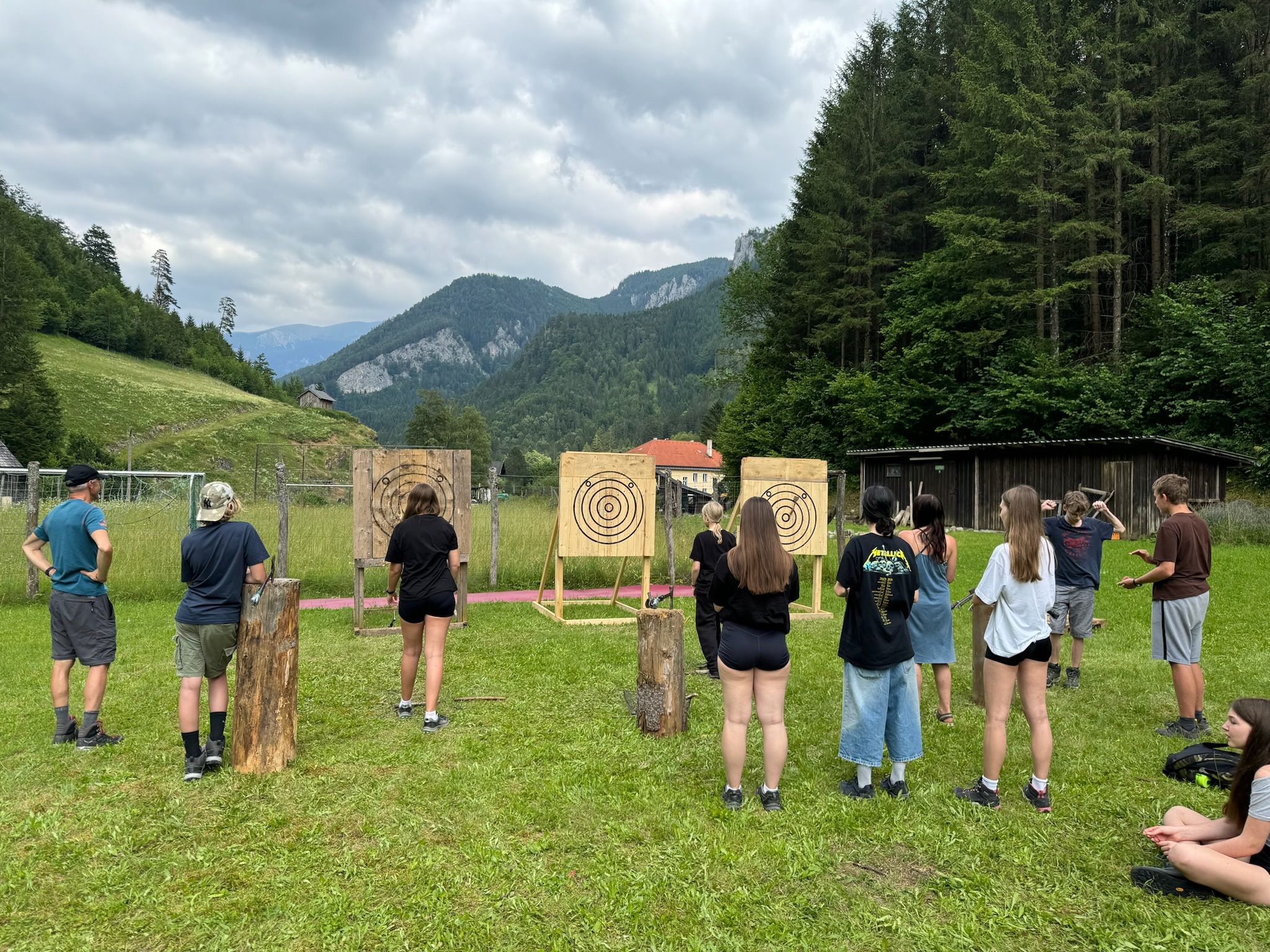 Group of people throwing axes outdoors in front of targets in a mountainous landscape.
