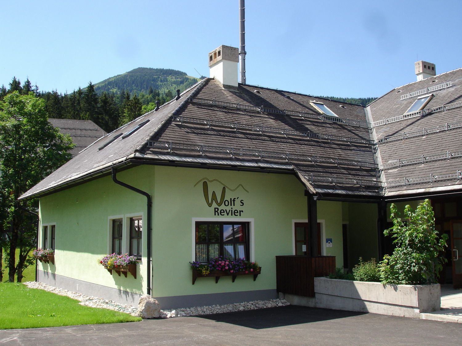 A green building with the inscription 'Wolf's Revier' and flower boxes in front of the windows, surrounded by trees and mountains in the background.
