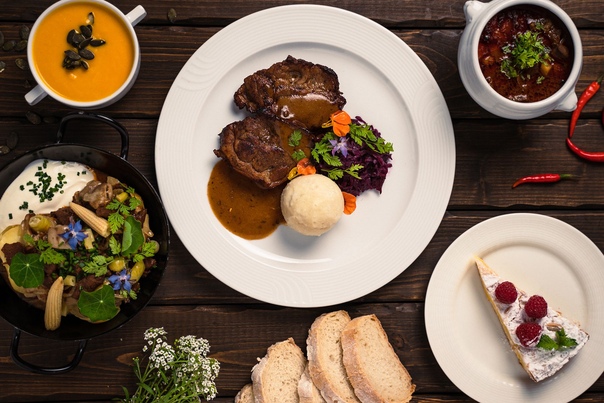 Various dishes on a wooden table, including soup, meat dish, stew, bread and cake.