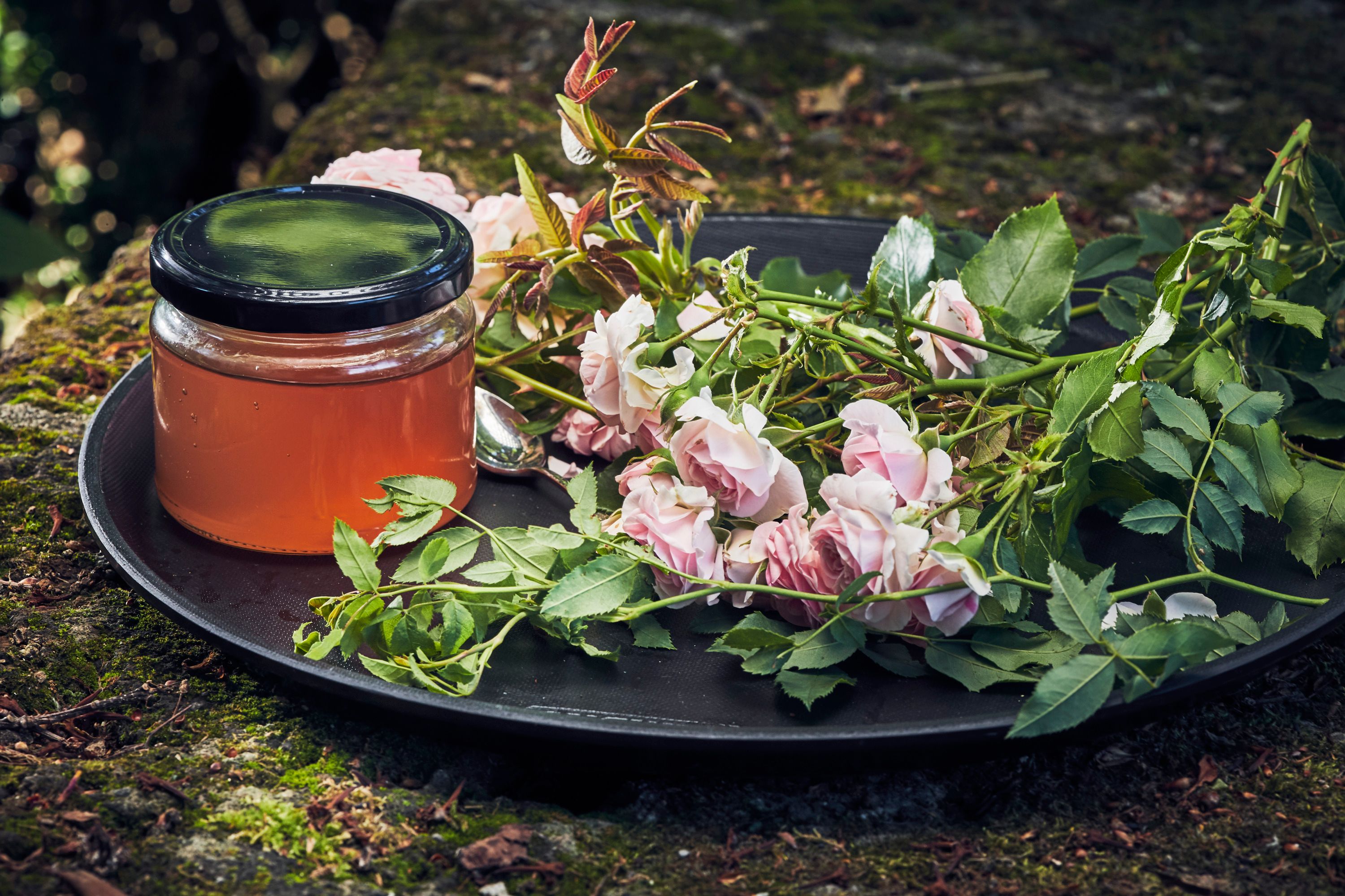 A glass of rose petal jelly next to fresh roses on a tray outside.