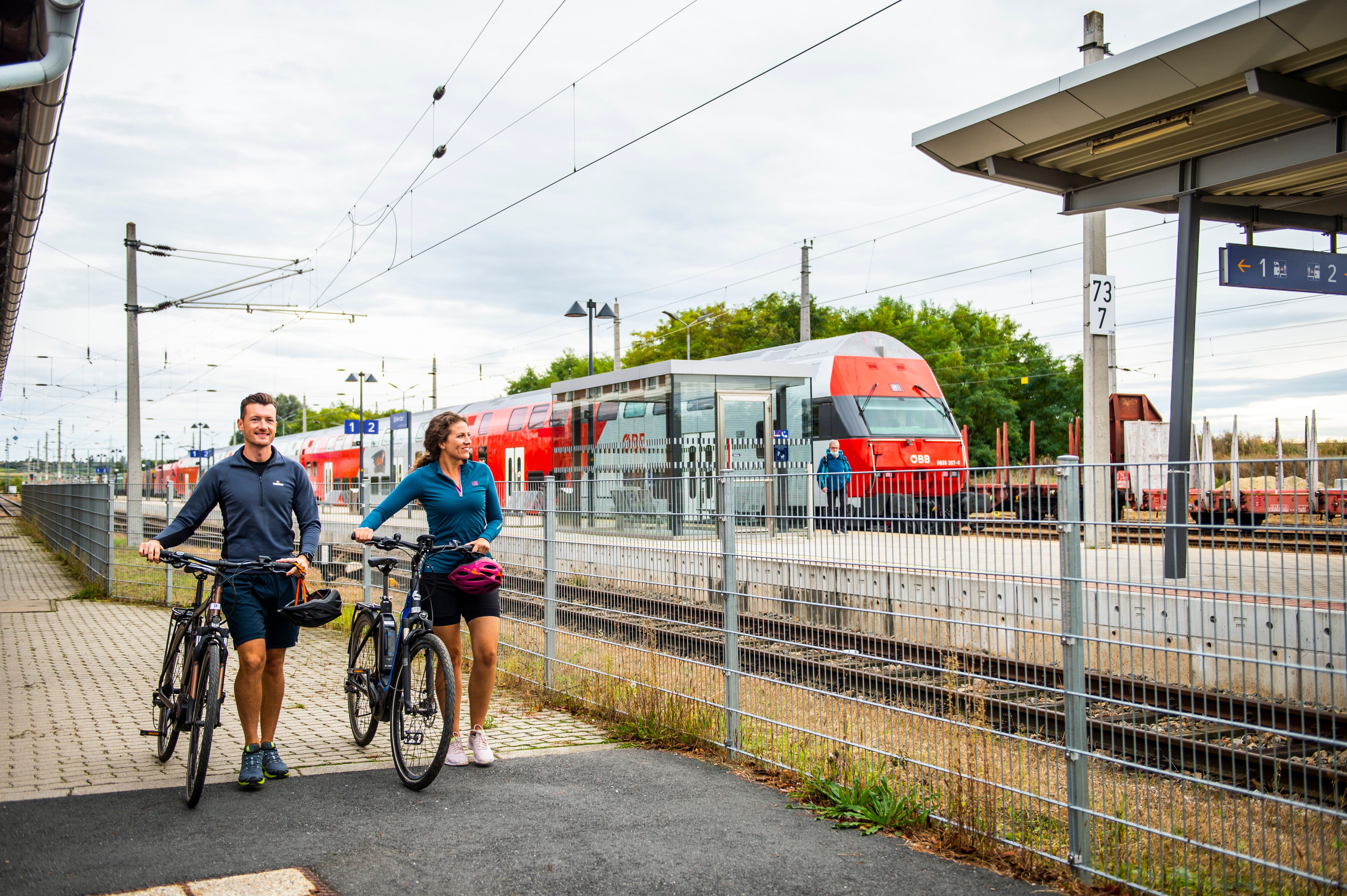 Two people with bicycles at the station, an ÖBB train in the background.