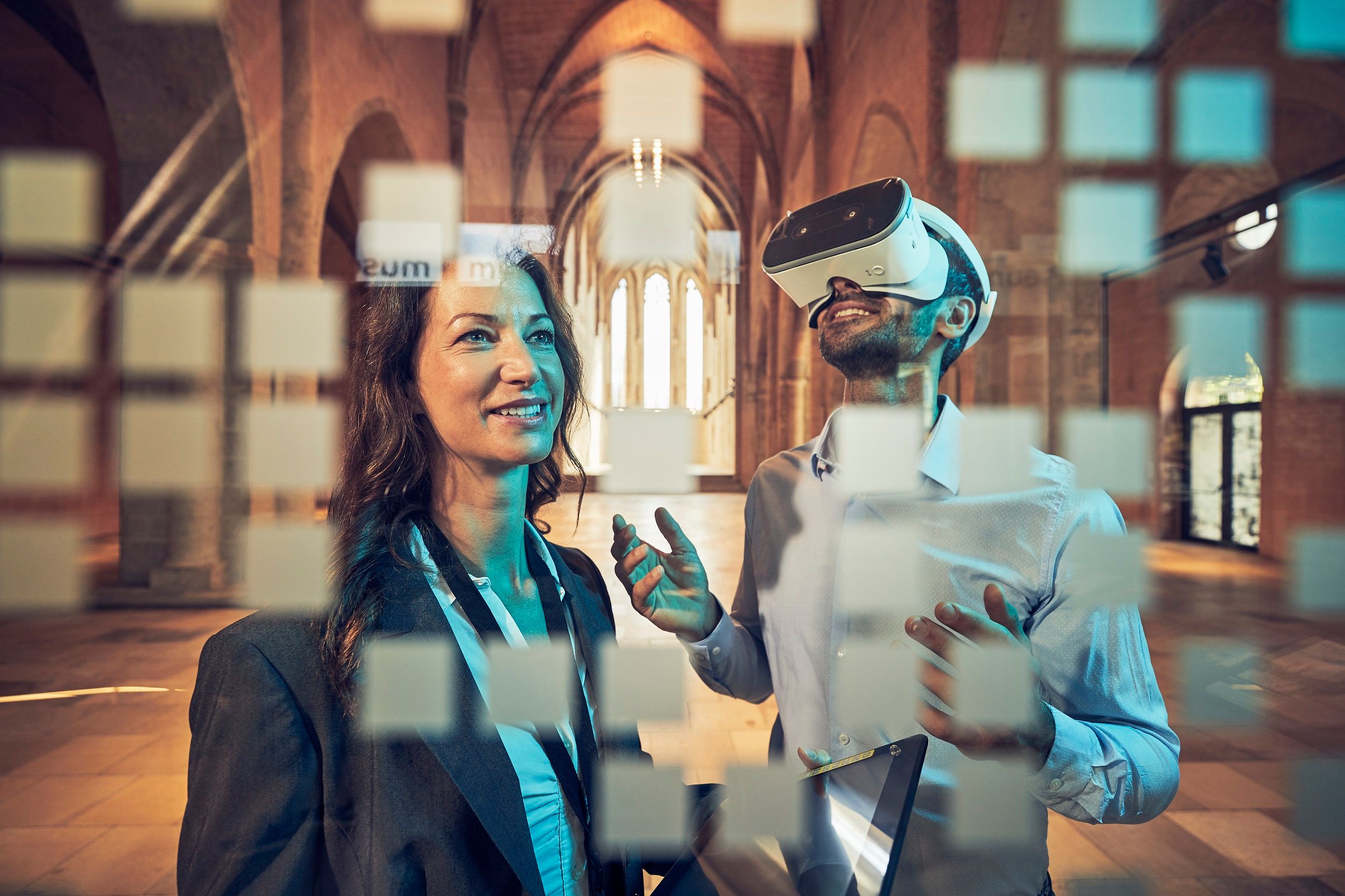 Two people in a historic building, one wearing VR glasses, the other smiling. Blurred squares can be seen in the foreground.