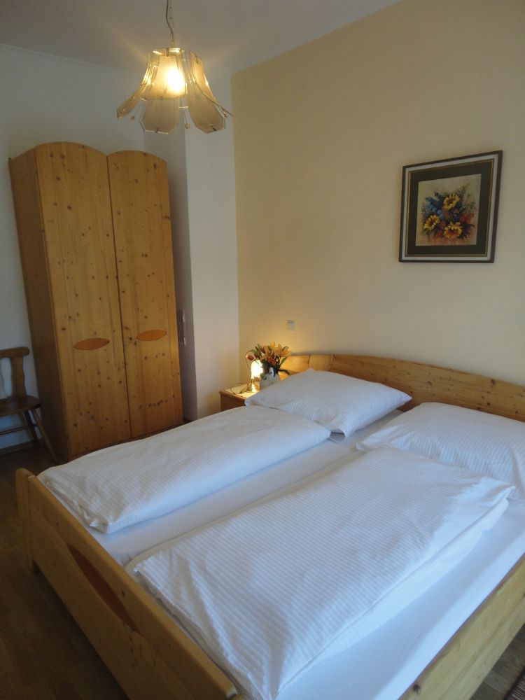 A cozy bedroom with double bed, wooden wardrobe, bedside table with flowers and a painting on the wall.