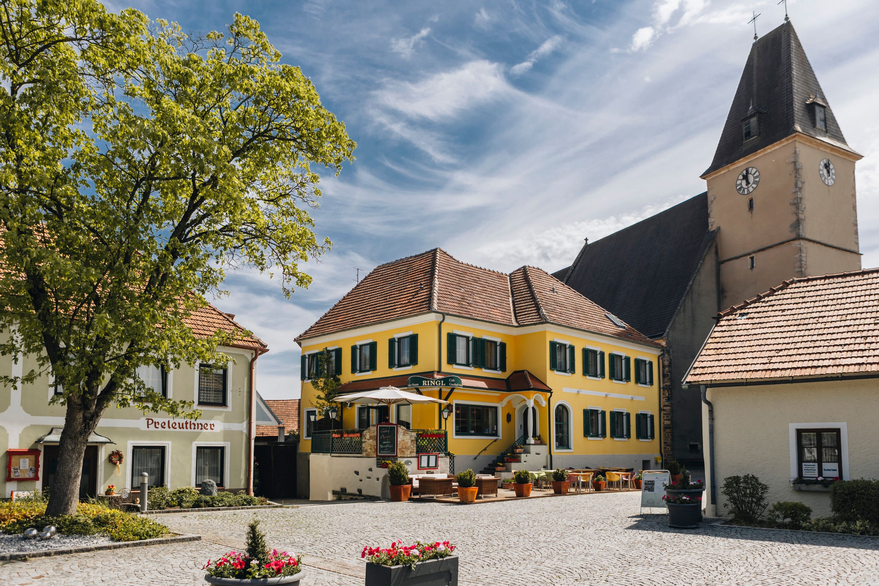 Idyllic village square with inn and terrace and a church tower in the background.