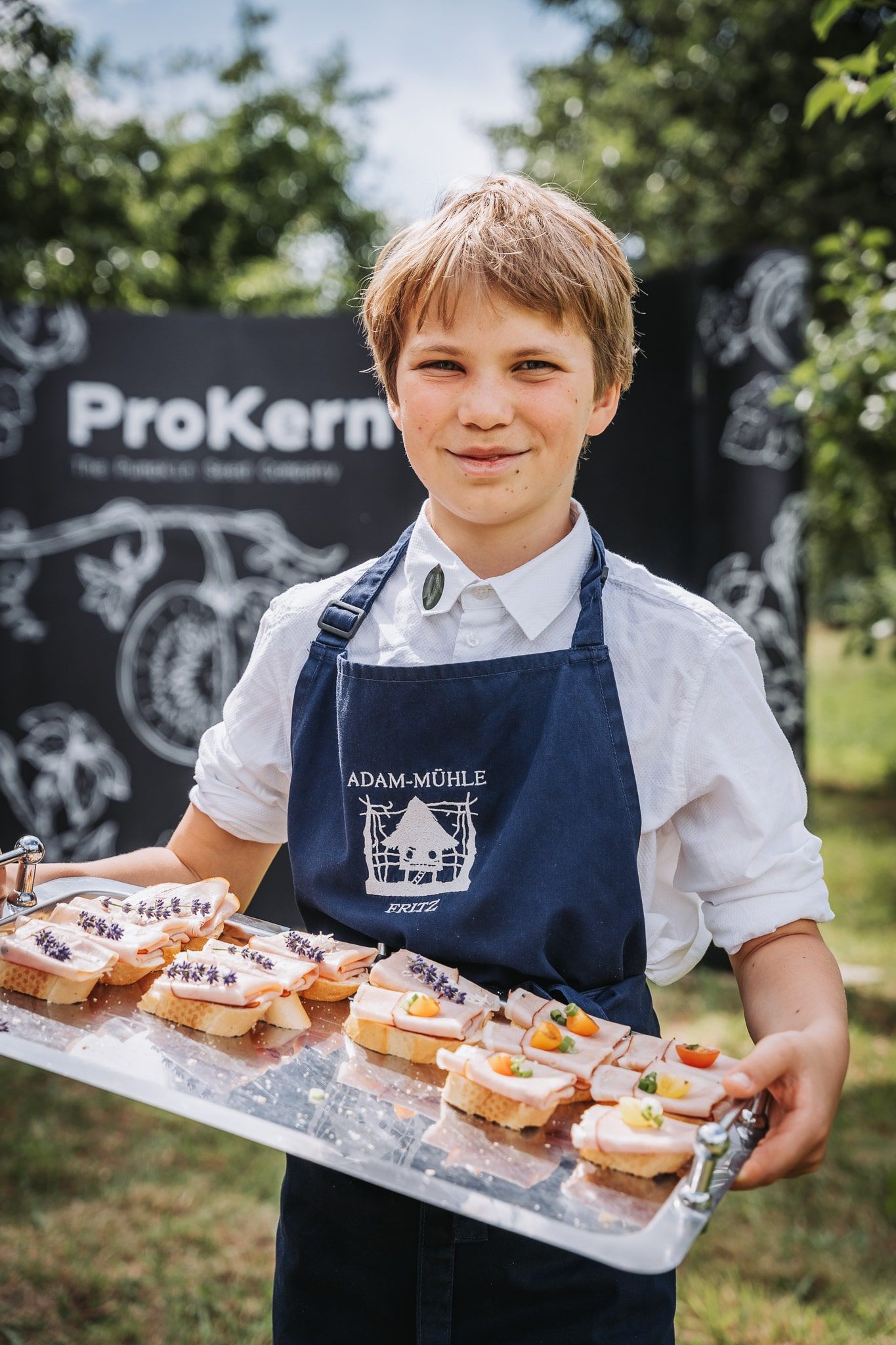 A child in a blue apron holds a tray of sandwiches outside.
