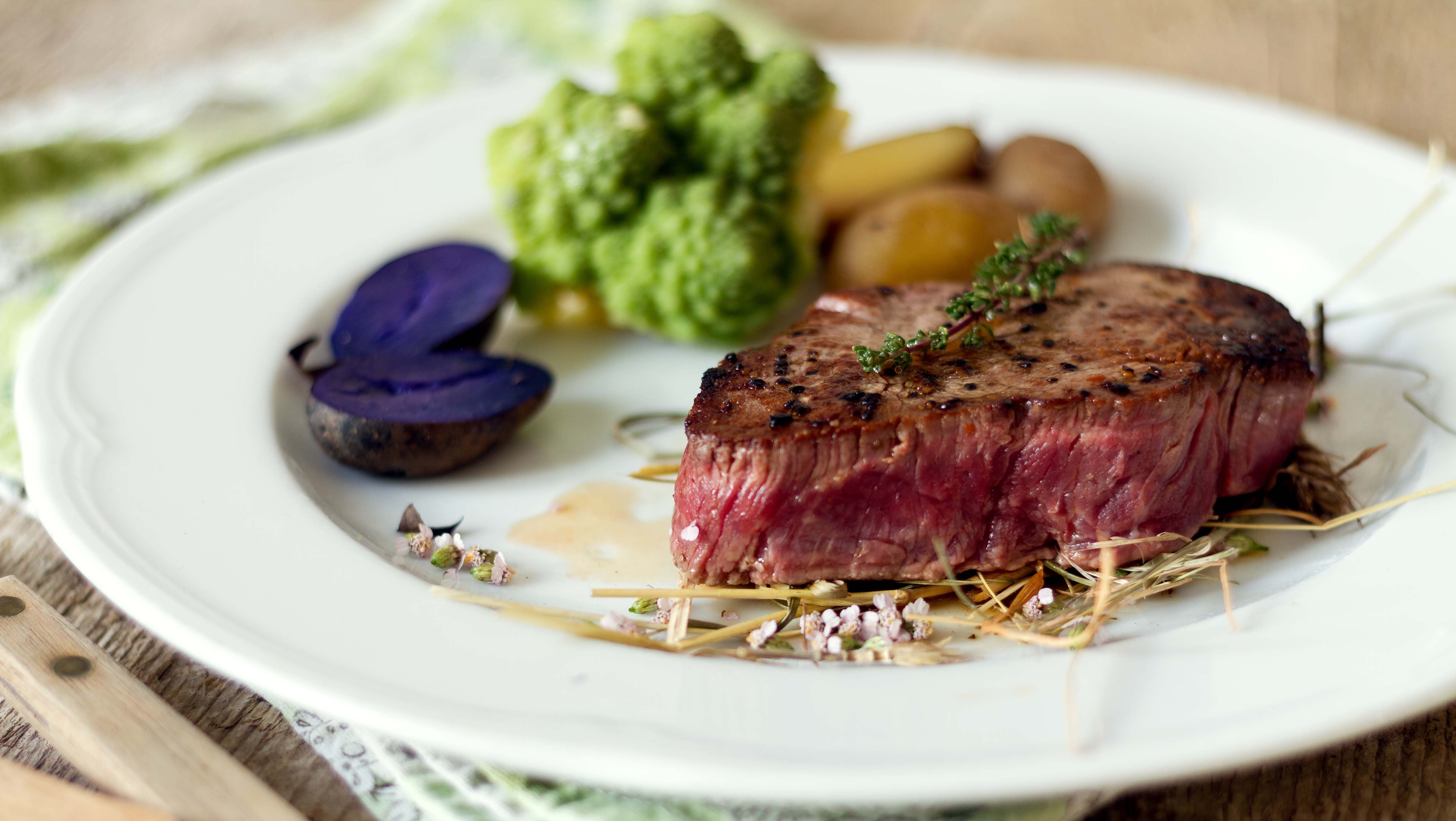 A piece of steak on a white round plate with vegetables, potatoes and herbs.