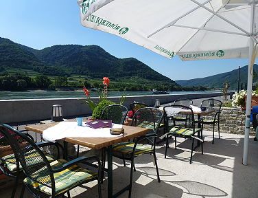 A guest garden with tables and chairs under parasols, with a view of the Danube and wooded hills in the background.