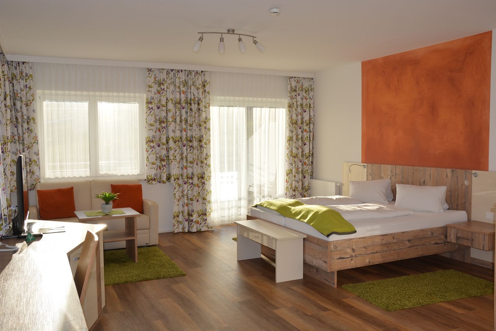 A modern double room with wooden floor, a double bed, sofa, table and large windows with curtains.