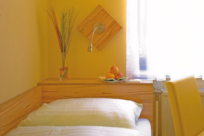 Cozy room with yellow walls, bed, vase with grasses and fruit bowl on a wooden shelf.