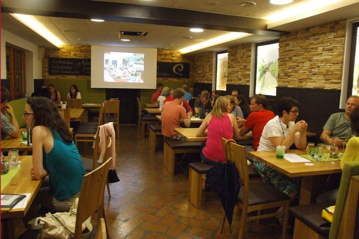 People sit at wooden tables in a cozy room while a presentation is shown on a screen.