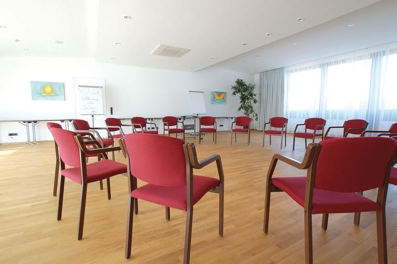 A bright seminar room with red chairs in a circle, a wooden floor and large windows.