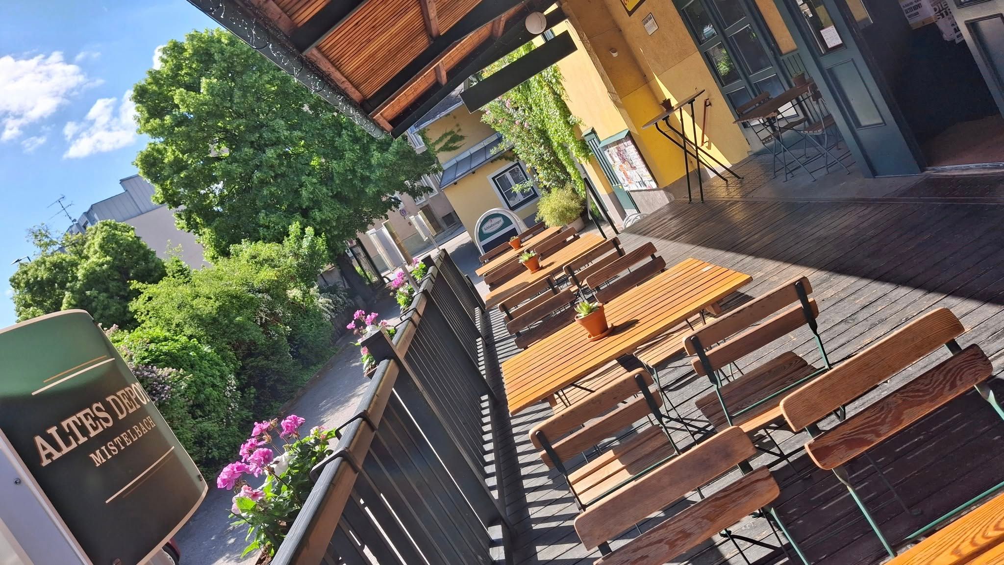A sunny terrace with wooden tables and chairs, surrounded by plants and a building with a yellow façade.
