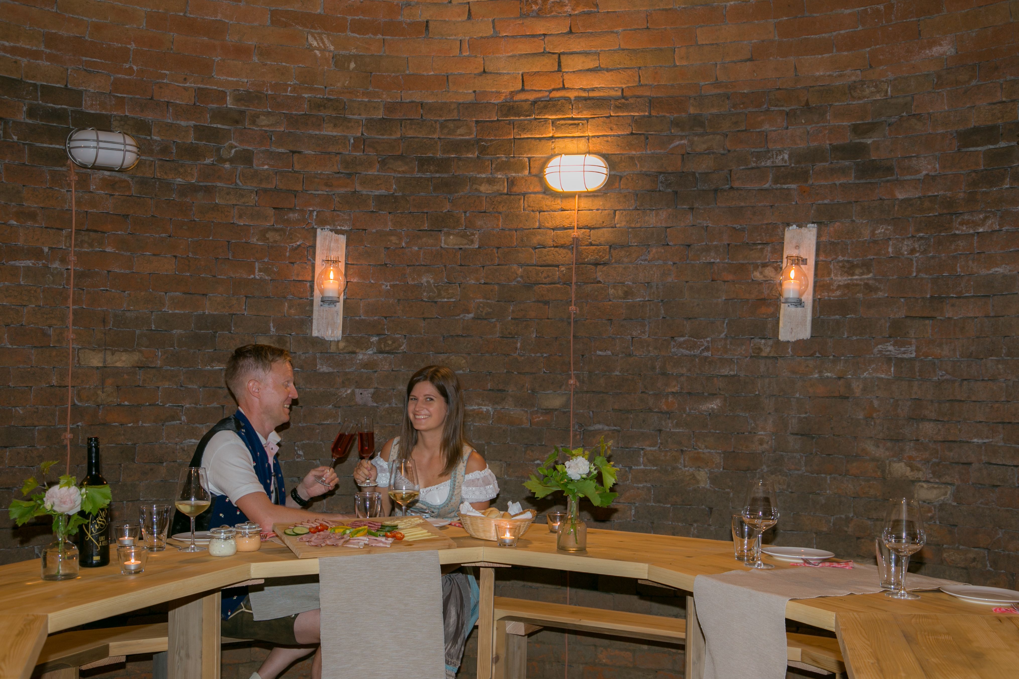 A couple sits at a wooden table in a wine cellar and toasts with red wine.