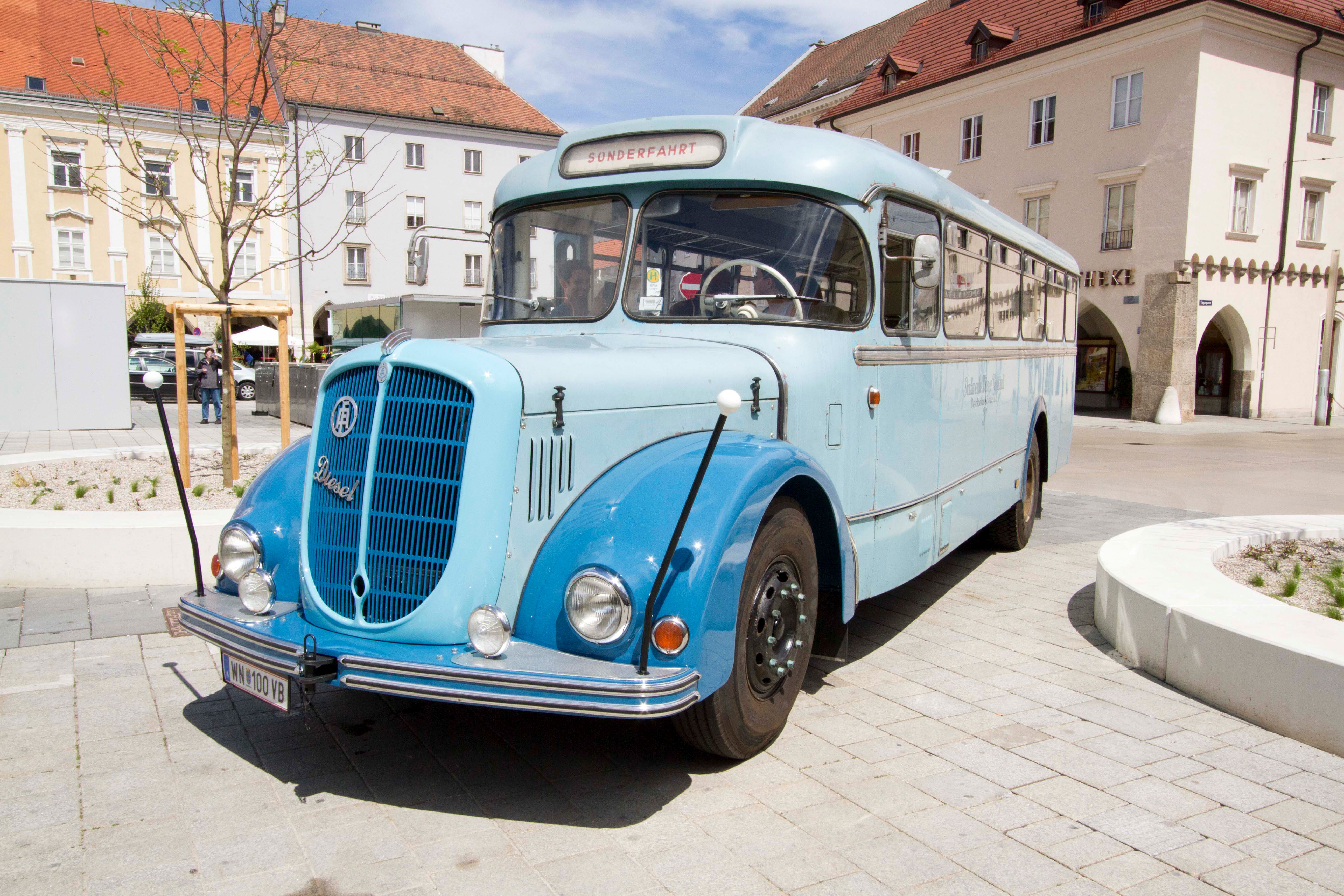 A blue vintage bus is parked on a cobbled square in front of historic buildings.