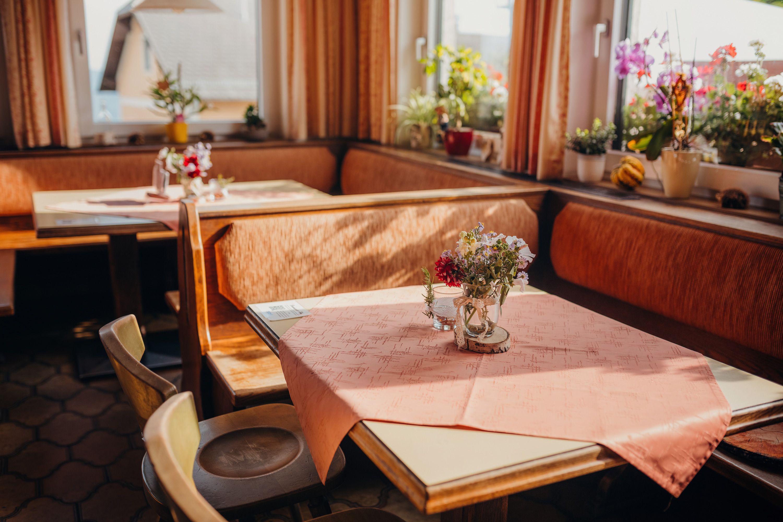 Cozy parlor with wooden furniture, tablecloths and flowers on the tables, surrounded by windows with plants.