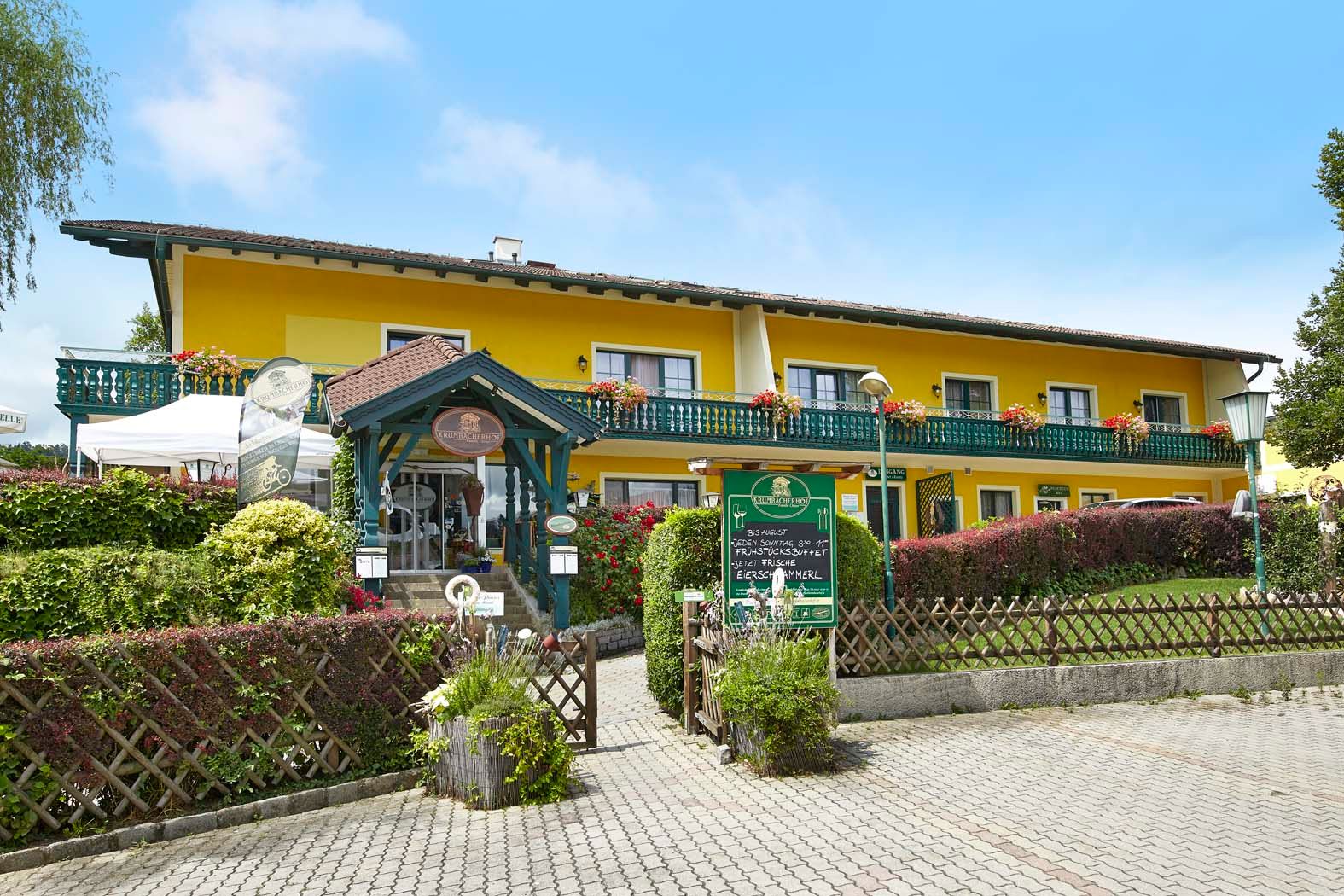 Yellow building with balcony and flowers, Krumbacherhof, entrance with canopy and signs.