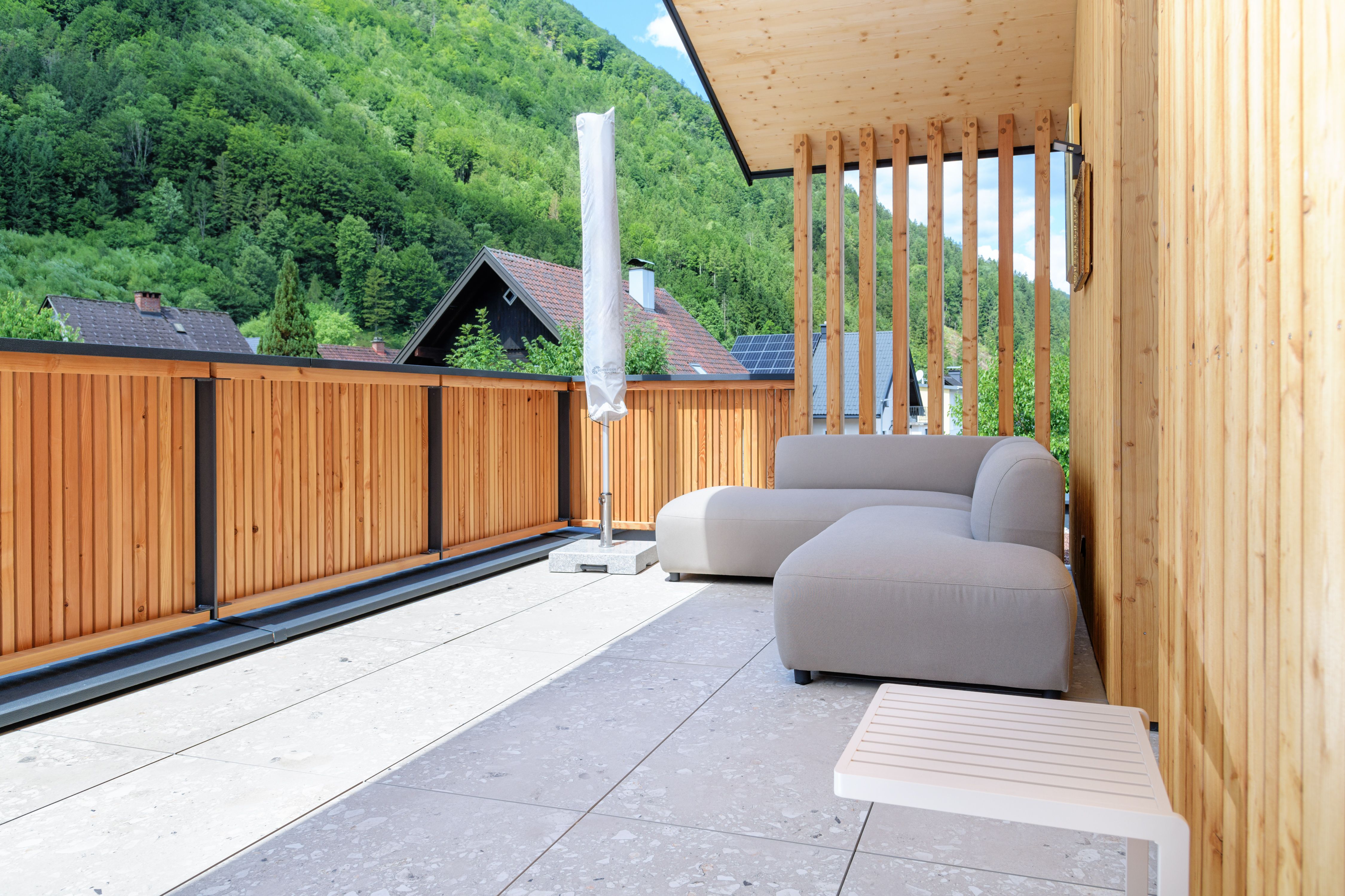 A modern patio with wooden walls, a gray sofa, and a view of wooded hills.