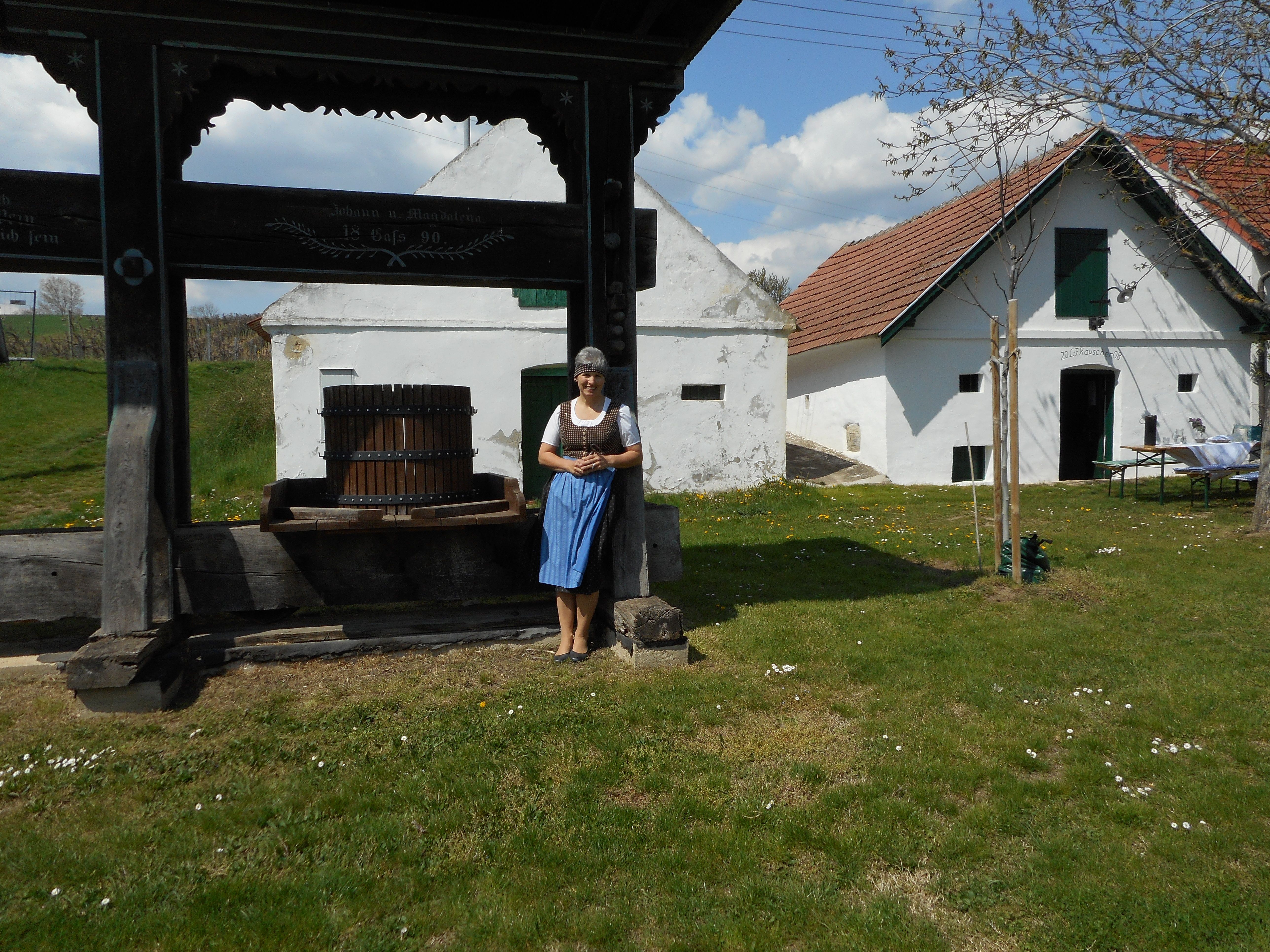 A woman in traditional dress stands next to an old wine press in front of a historic building with white walls and red roofs.