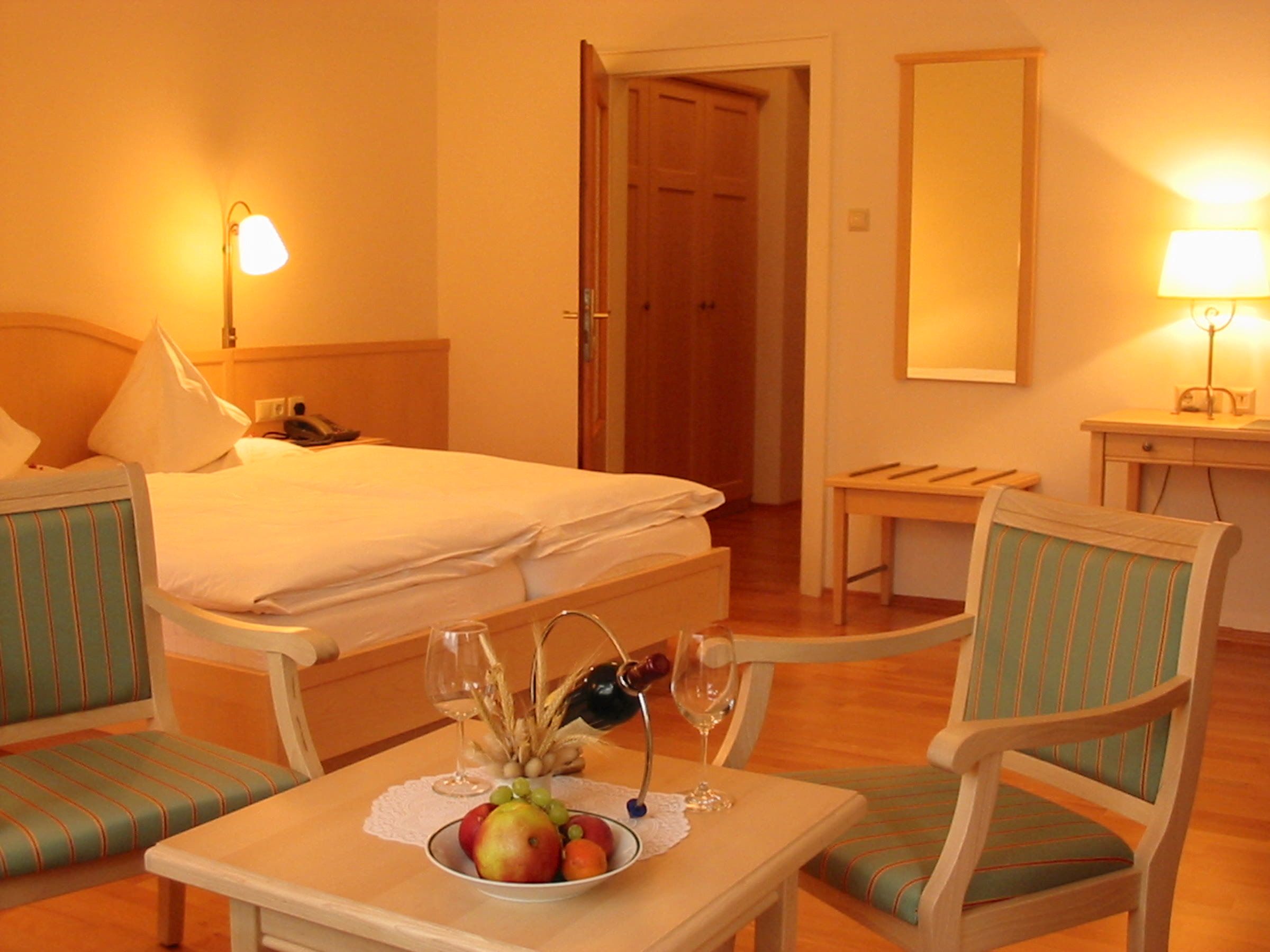 A cozy hotel room with a double bed, two chairs, a table with fruit and wine, and warm light.