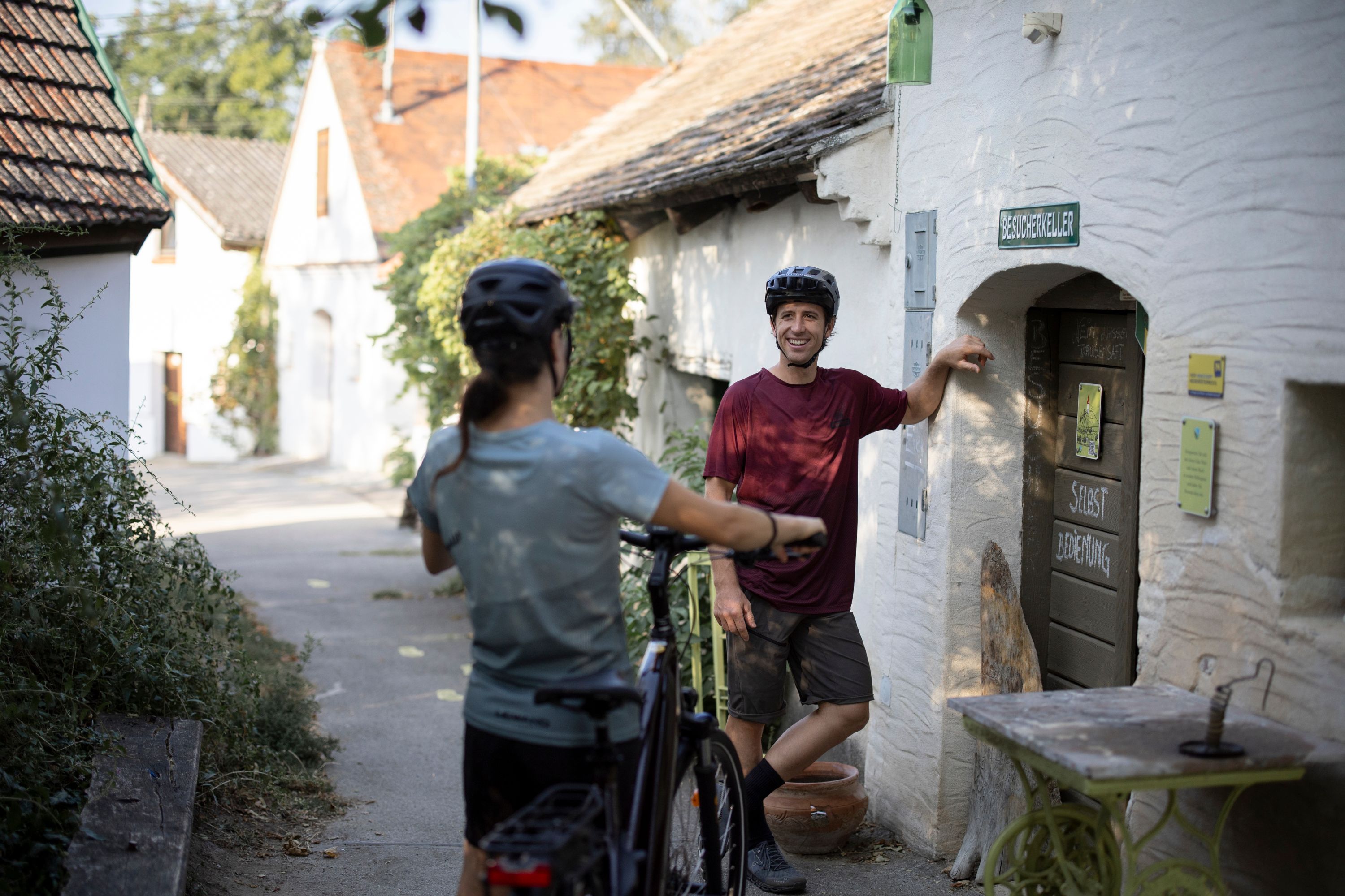 Two cyclists in a wine cellar lane, one leaning against a door labeled 'Besucherkeller'.