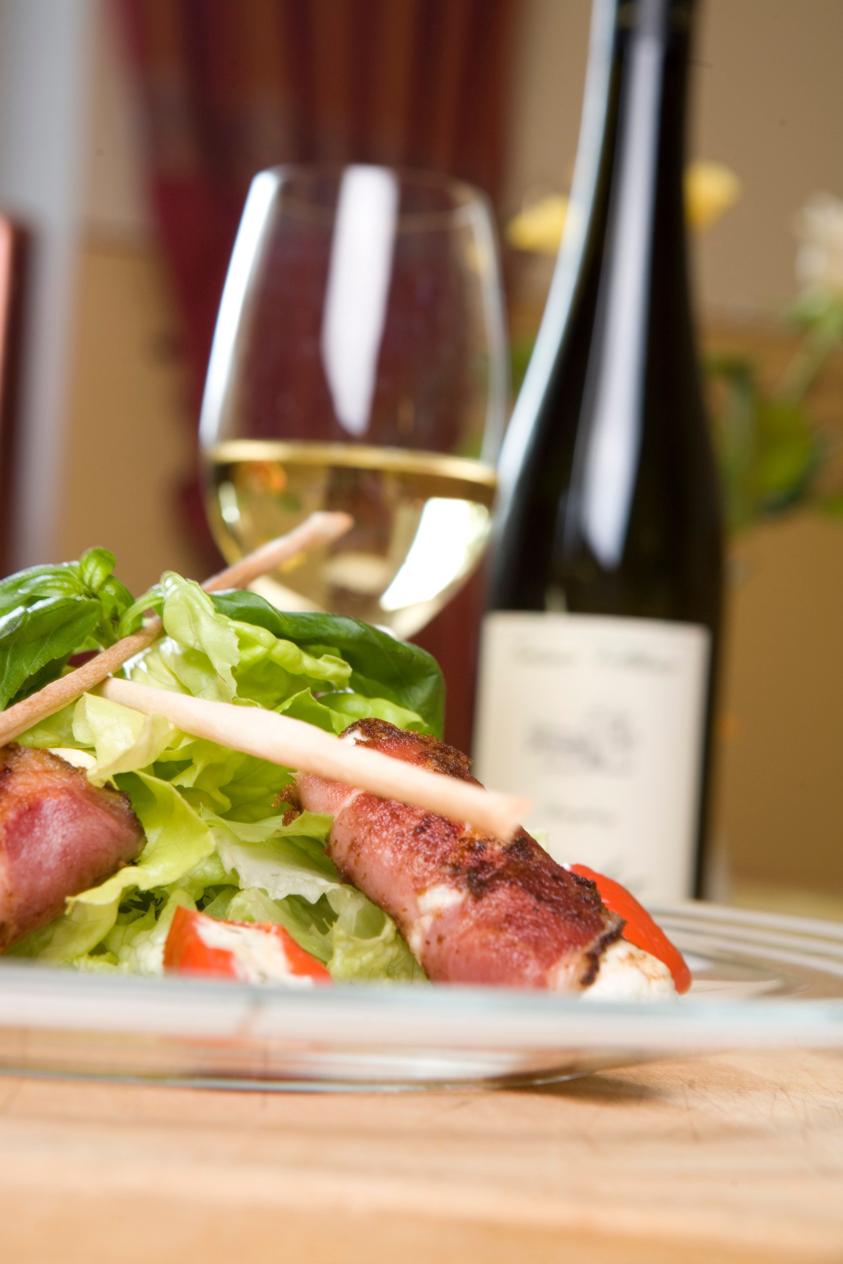 A plate with salad and cheese wrapped in bacon, a wine glass and a bottle of wine in the background.