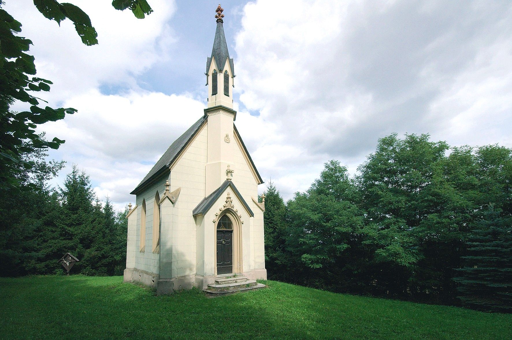 A small chapel with a pointed tower, surrounded by trees and green lawns, under a cloudy sky.