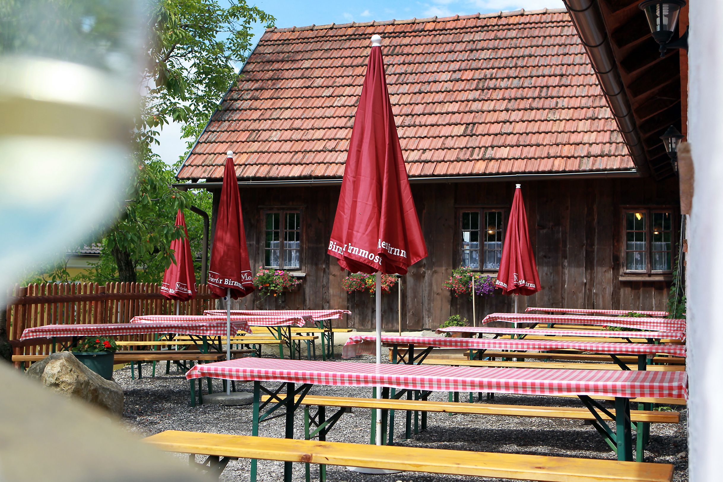 A traditional wine tavern with wooden tables, red and white checkered tablecloths and red umbrellas in front of a rustic building with a tiled roof.