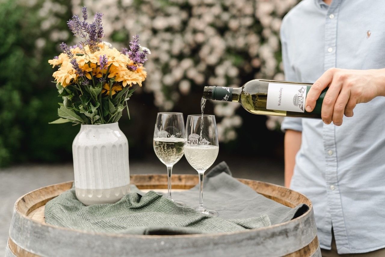A person is pouring white wine into two glasses on a wine barrel, with a vase of flowers next to it.