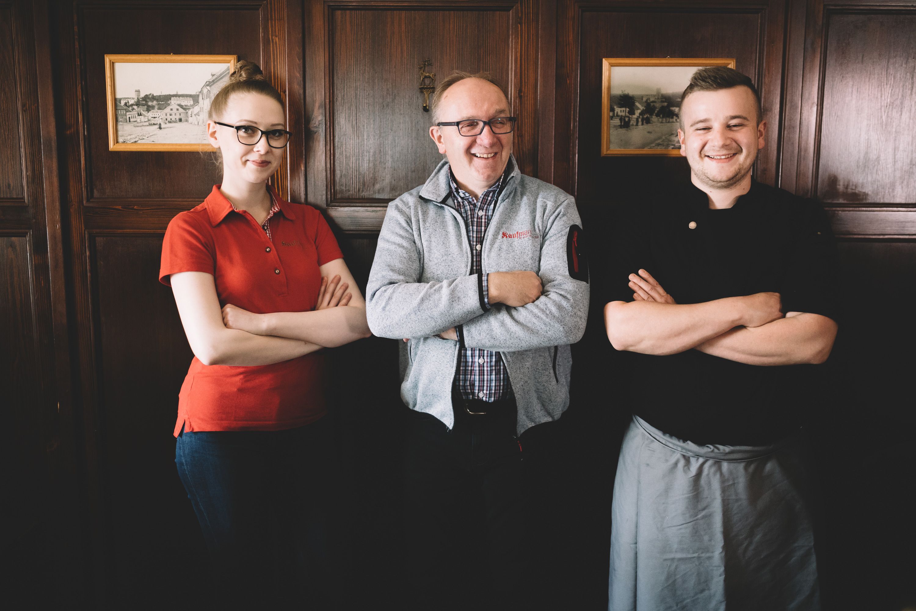 Three people stand smiling with their arms folded in front of a wooden wall.
